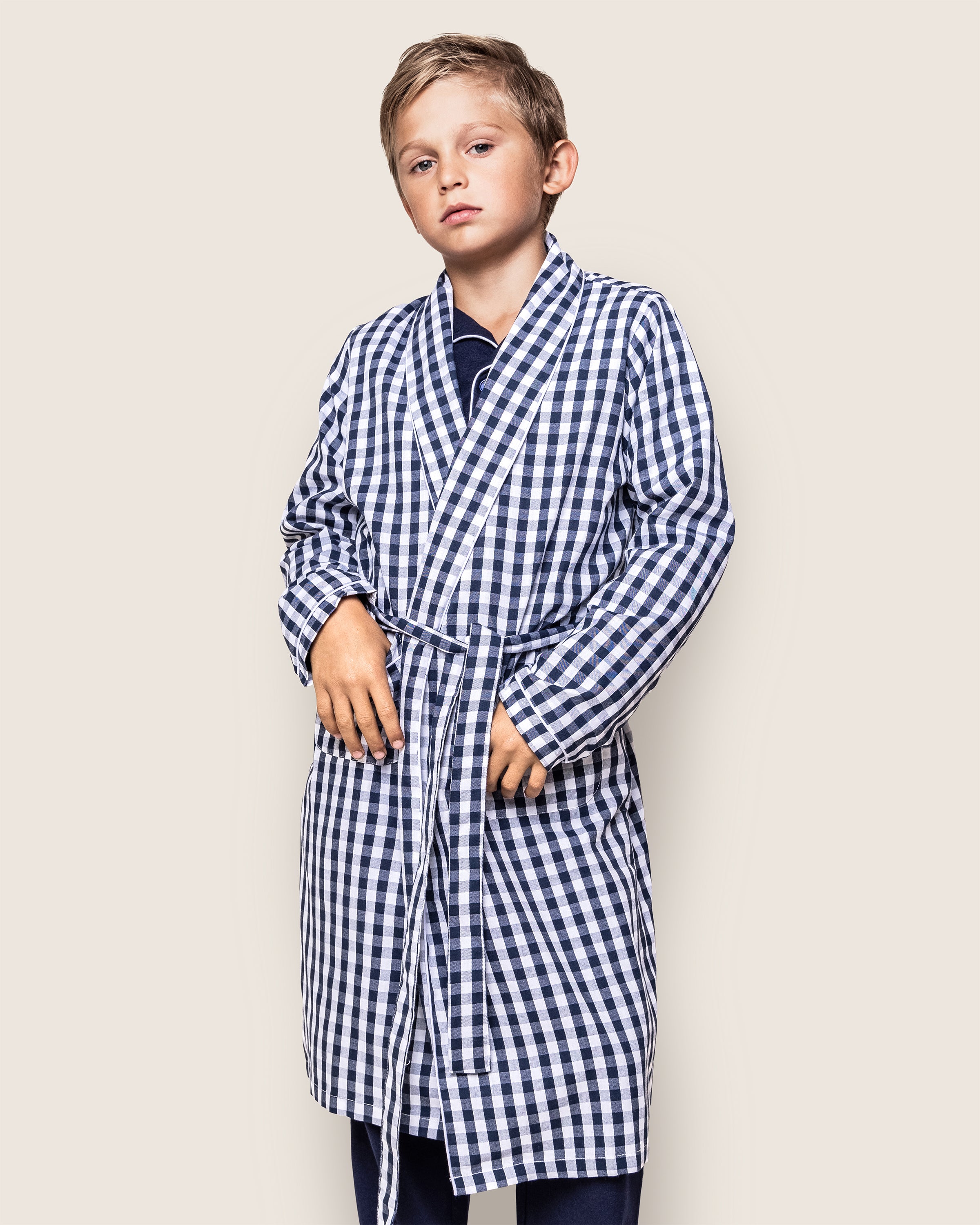 A young boy wearing Petite Plumes Kids Twill Robe in Navy Gingham over a dark shirt stands against a neutral background, holding the robes belt. With a relaxed gaze at the camera, he embodies both style and comfort in childrens sleepwear.