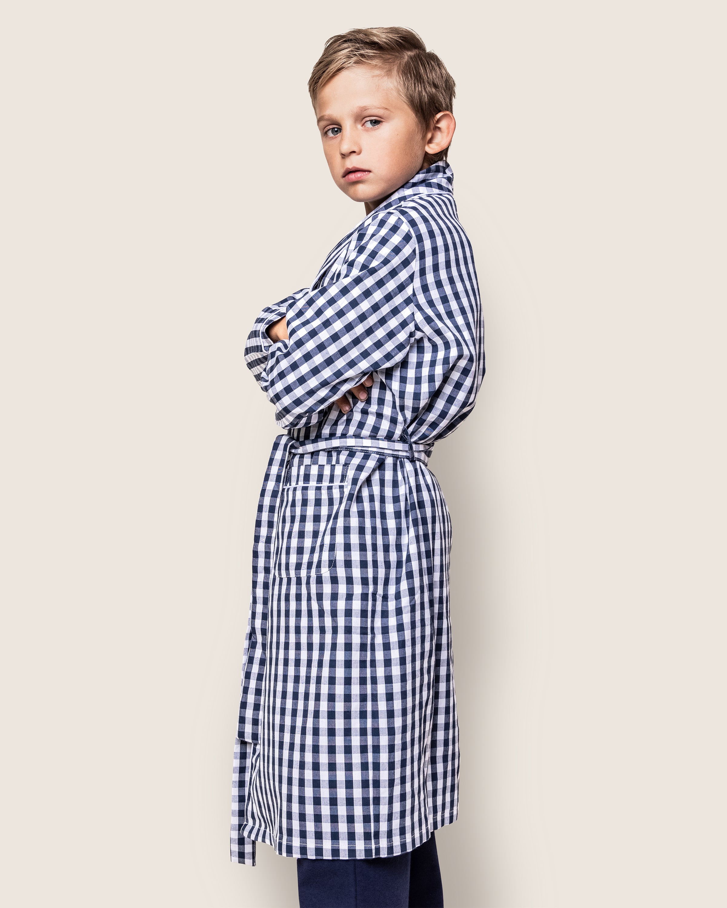 A young boy wearing a navy gingham Kids Twill Robe by Petite Plume, crafted from flame-retardant fabric, stands with arms crossed, looking over his shoulder against a plain background.