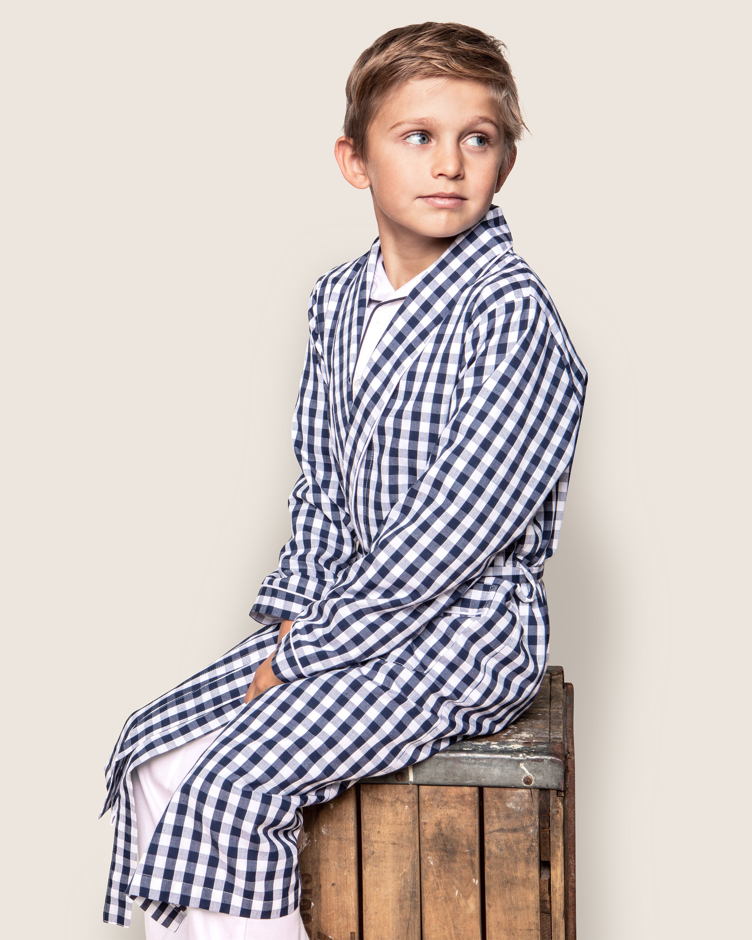 A child in a Petite Plume Kids Twill Robe in Navy Gingham sits on a wooden crate against an off-white backdrop, short hair framing their thoughtful expression, capturing a serene moment in delightful childrens sleepwear.