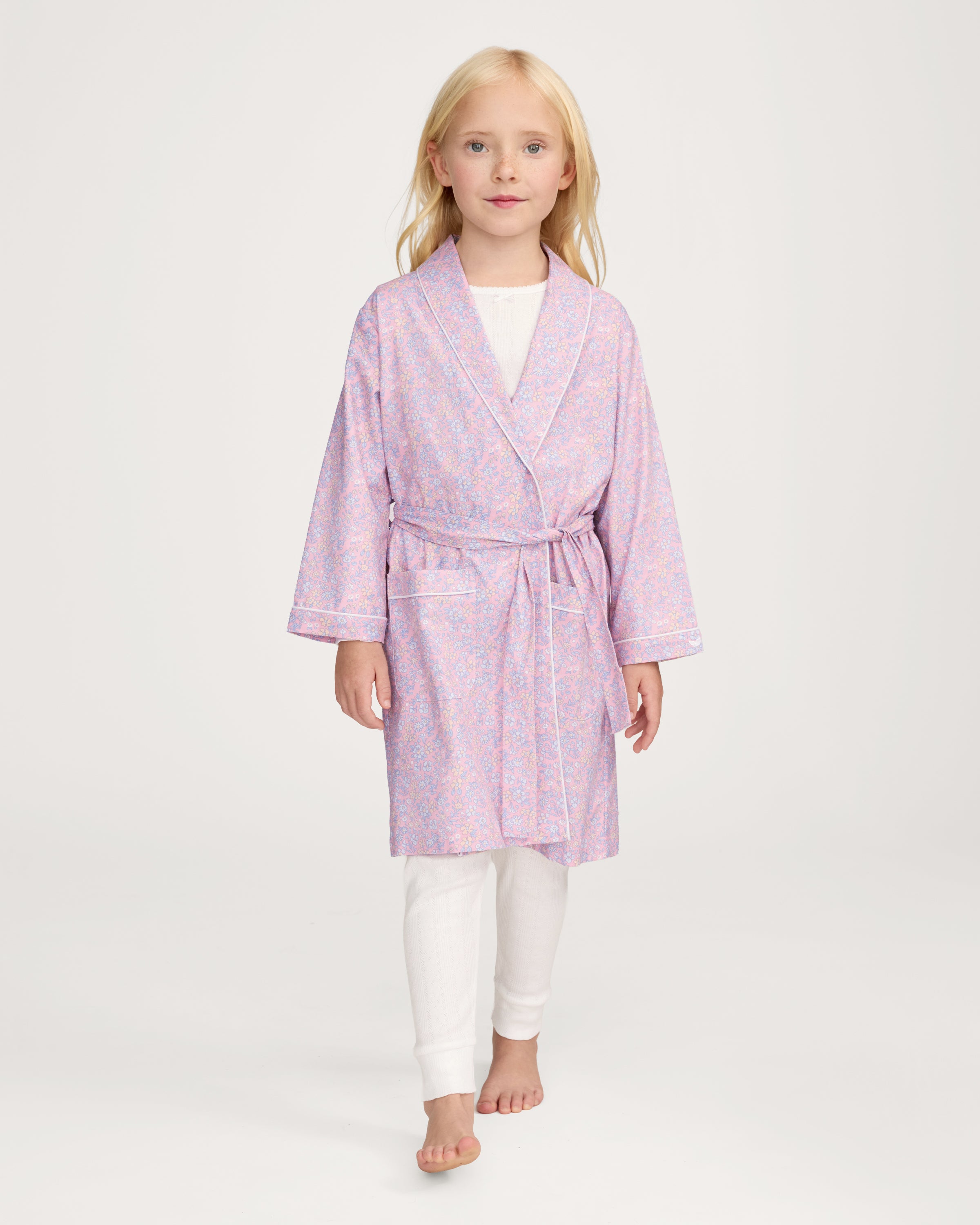 A young girl with blonde hair smiles as she ties the belt on her Kid's Twill Robe in Fleur Blush over a white nightgown.