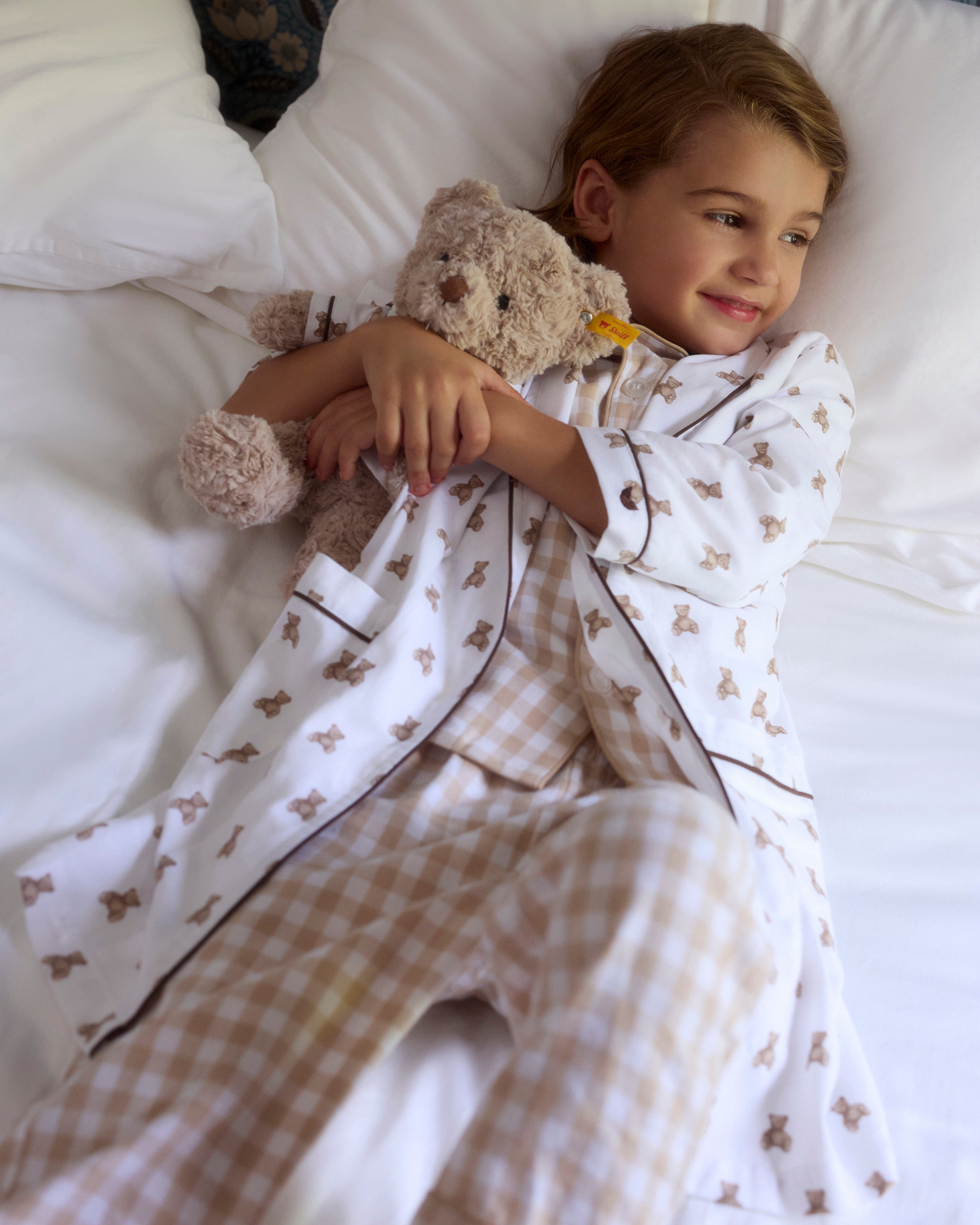 The Petite Plume x Steiff Teddy Bear, a plush light brown bear in a white shirt with teddy bear prints and a yellow ear tag, sits on a bed against a dark brown pillow.