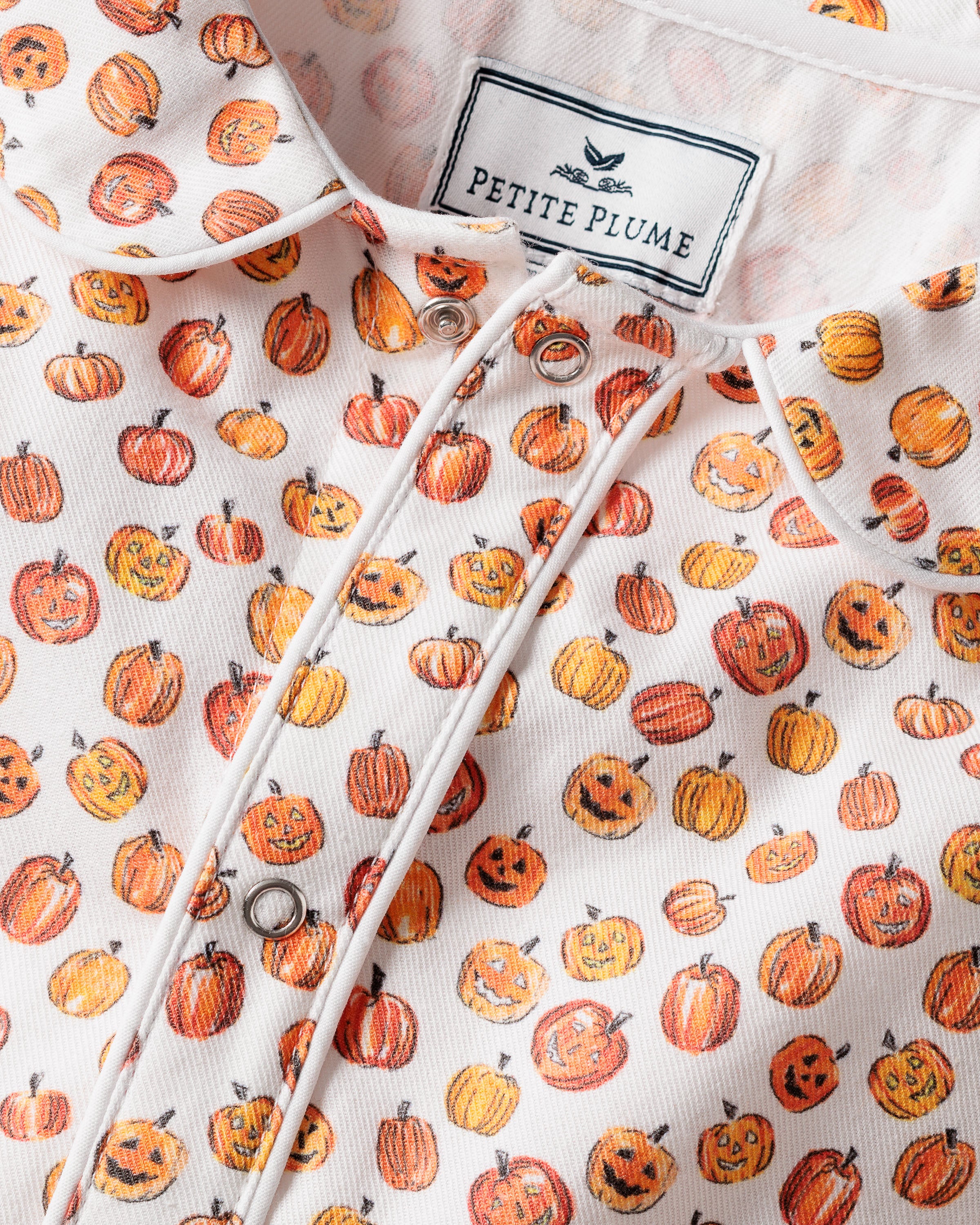 Close-up of the Babys Twill Cambridge Romper in Pumpkin Patch by Petite Plume, featuring a white fabric with friendly orange jack-o-lanterns. The romper is accented with metal snap buttons and a Petite Plume tag at the top.