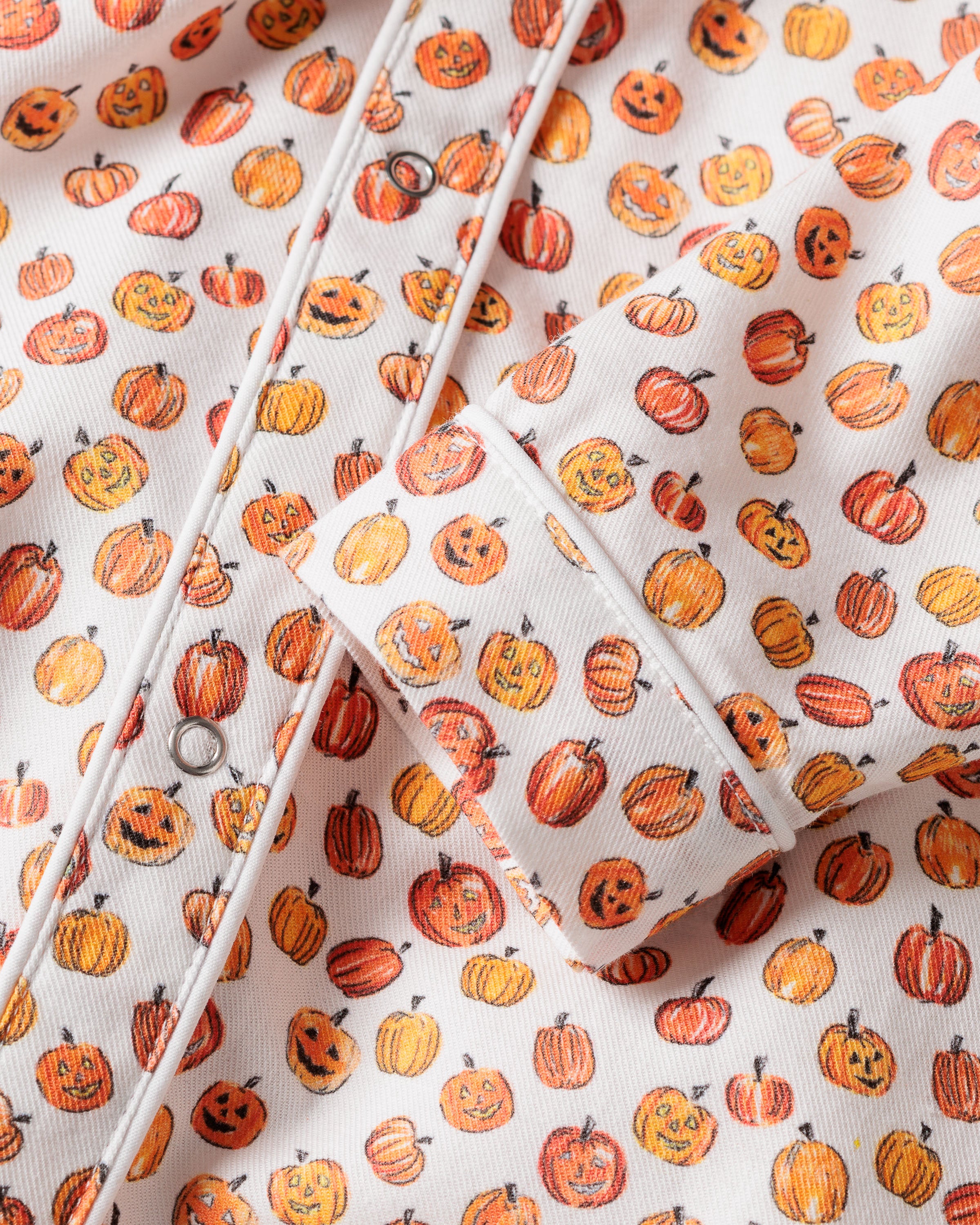 Close-up of a playful Halloween-themed fabric from the Babys Twill Cambridge Romper in Pumpkin Patch by Petite Plume, featuring kid-friendly prints of festive pumpkins and jack-o-lanterns on white. The garment includes visible white piping and buttons.