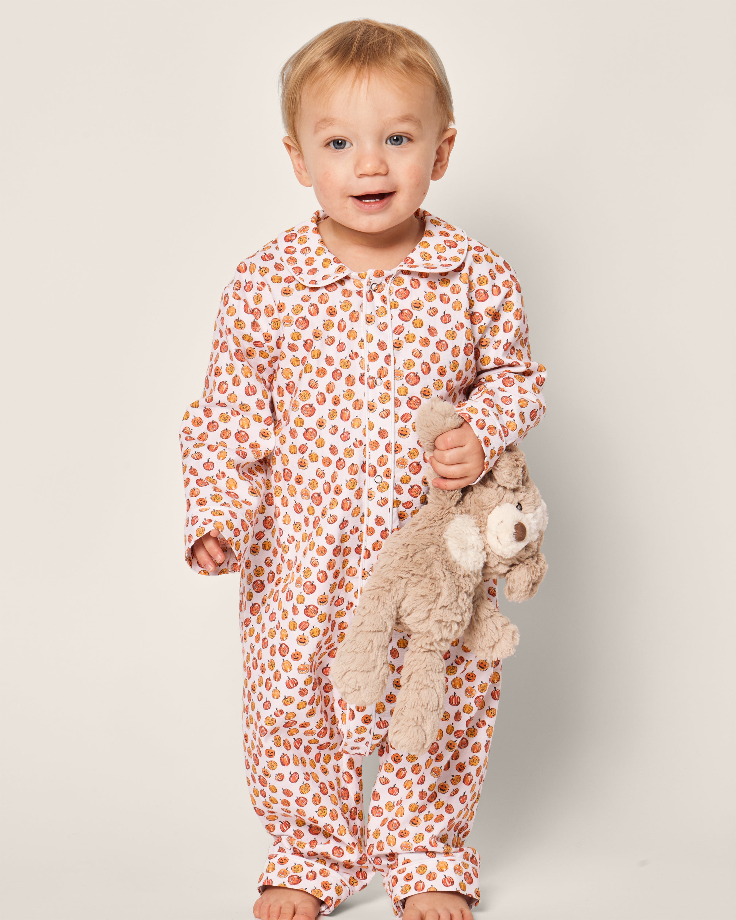 A toddler in Petite Plumes Babys Twill Cambridge Romper in Pumpkin Patch stands barefoot on a light background, holding a beige stuffed animal.