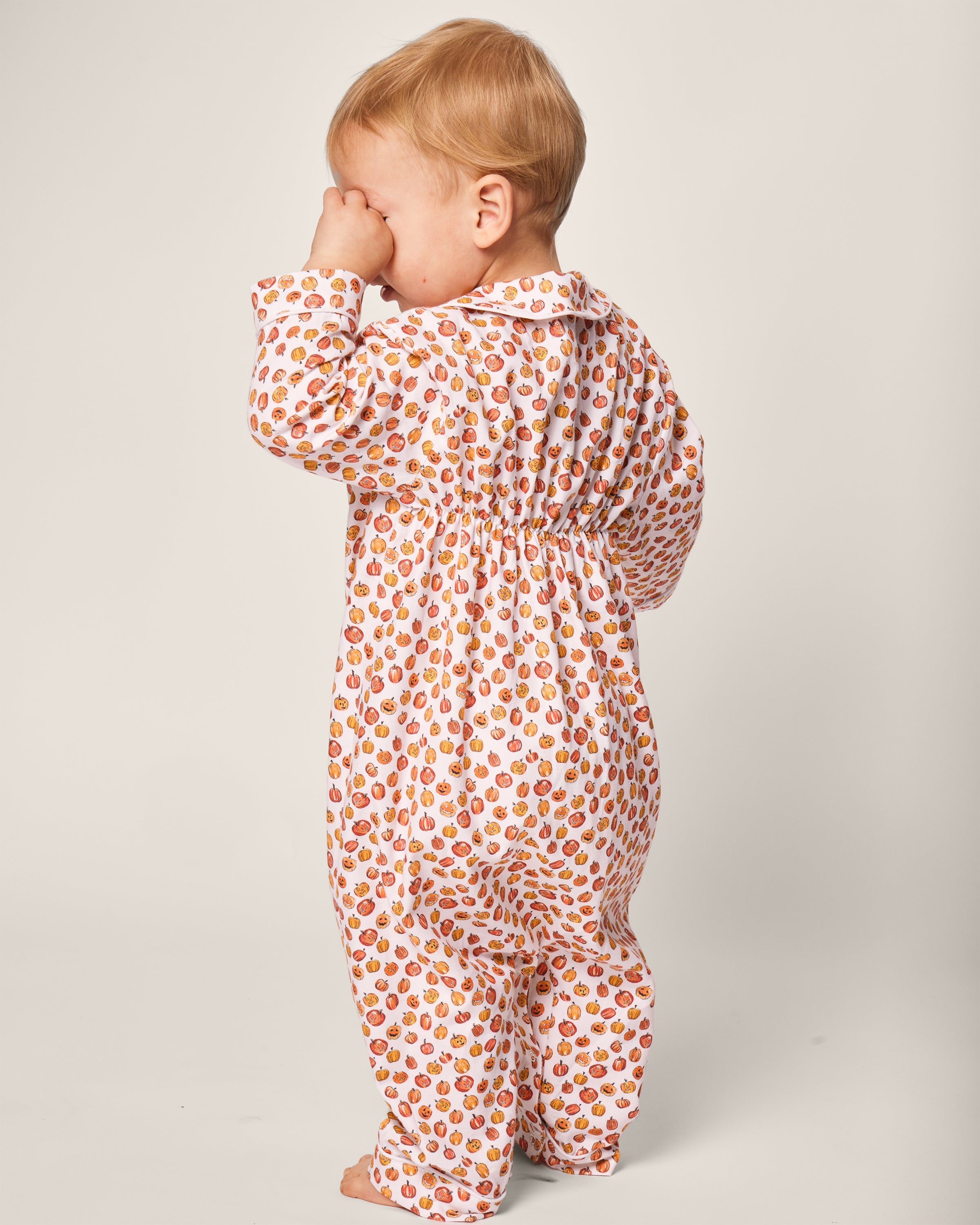 A toddler faces away in Petite Plumes Babys Twill Cambridge Romper in Pumpkin Patch, a patterned onesie with small orange and red shapes like festive pumpkins. The child rubs their face with one hand against a plain, light-colored background.