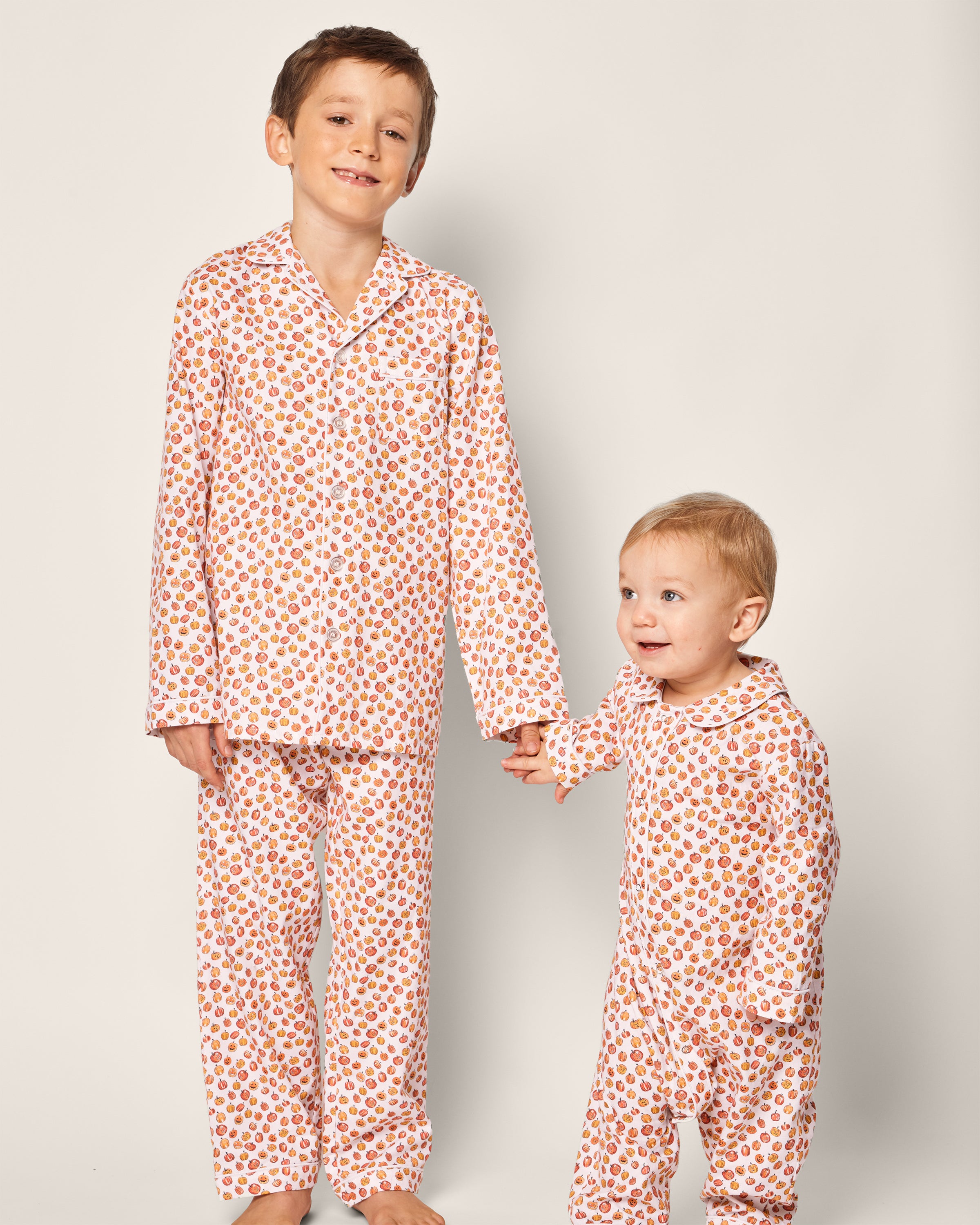 Two young children in matching Petite Plume pajamas, featuring playful Halloween prints from the Babys Twill Cambridge Romper in Pumpkin Patch line, hold hands. The older child stands smiling to the left against a plain light background.