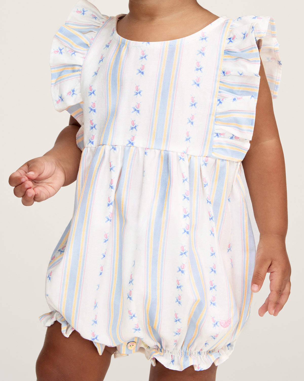 A smiling toddler with curly hair stands barefoot against a plain white background, wearing the Baby's Twill Ruffled Romper in Cottage Stripe—featuring pastel stripes, small floral patterns, and ruffled sleeves.