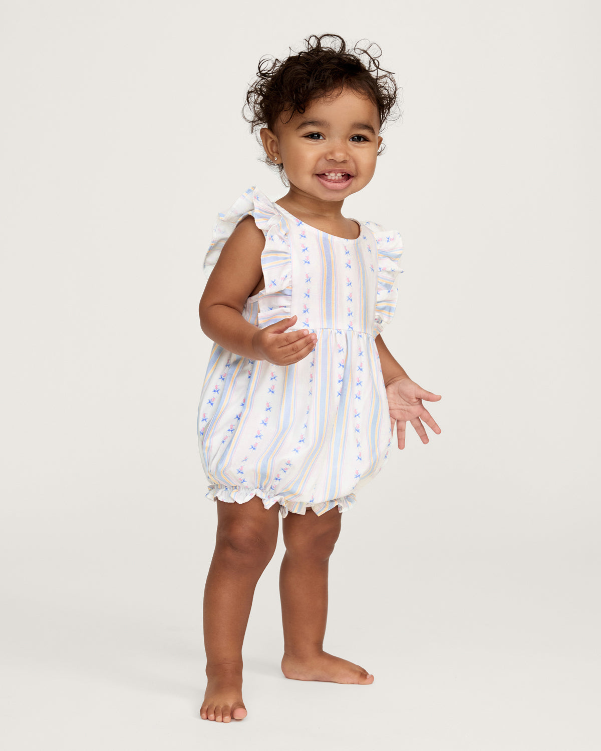 A smiling toddler with curly hair stands barefoot against a plain white background, wearing the Baby's Twill Ruffled Romper in Cottage Stripe—featuring pastel stripes, small floral patterns, and ruffled sleeves.
