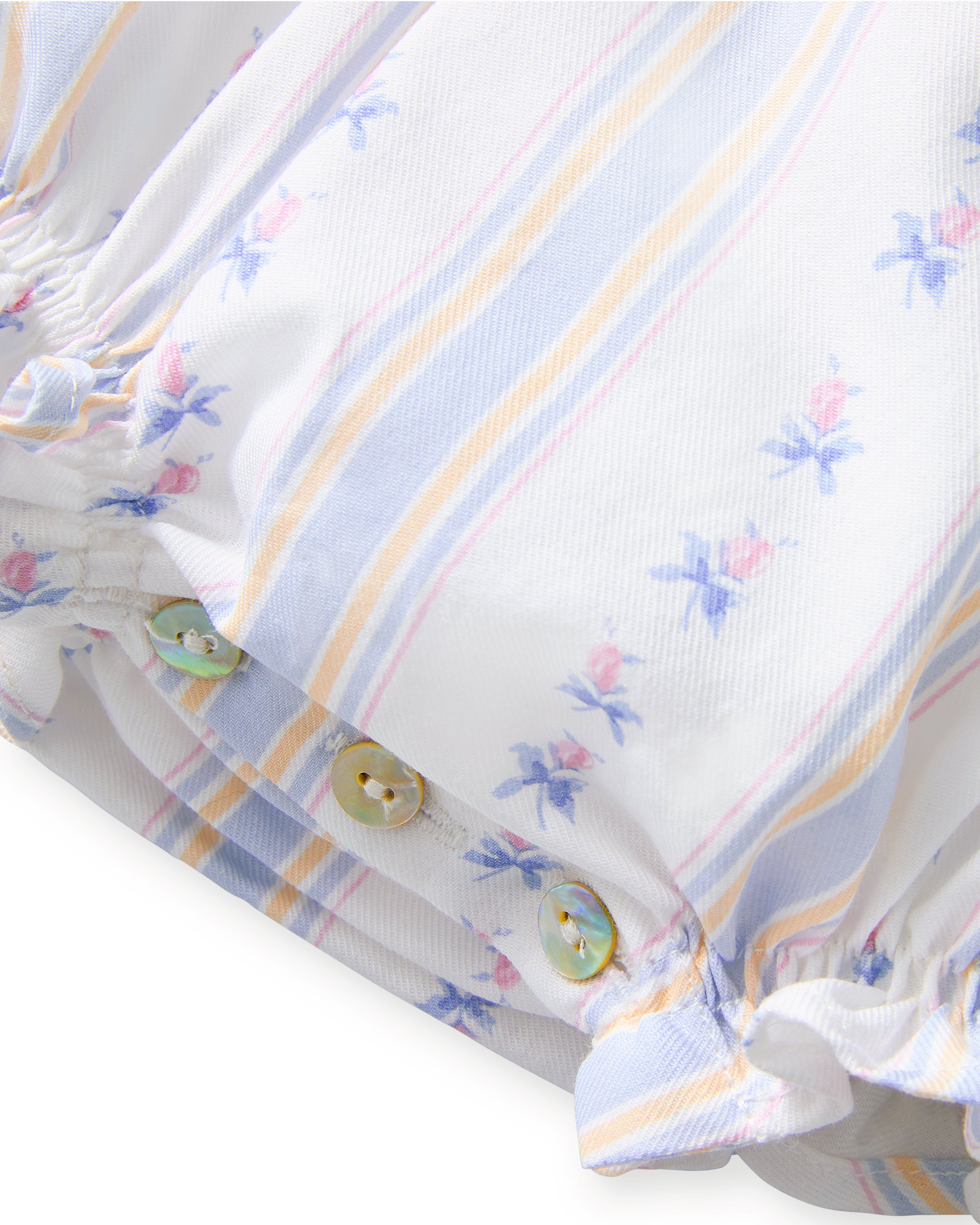 A smiling toddler with curly hair stands barefoot against a plain white background, wearing the Baby's Twill Ruffled Romper in Cottage Stripe—featuring pastel stripes, small floral patterns, and ruffled sleeves.