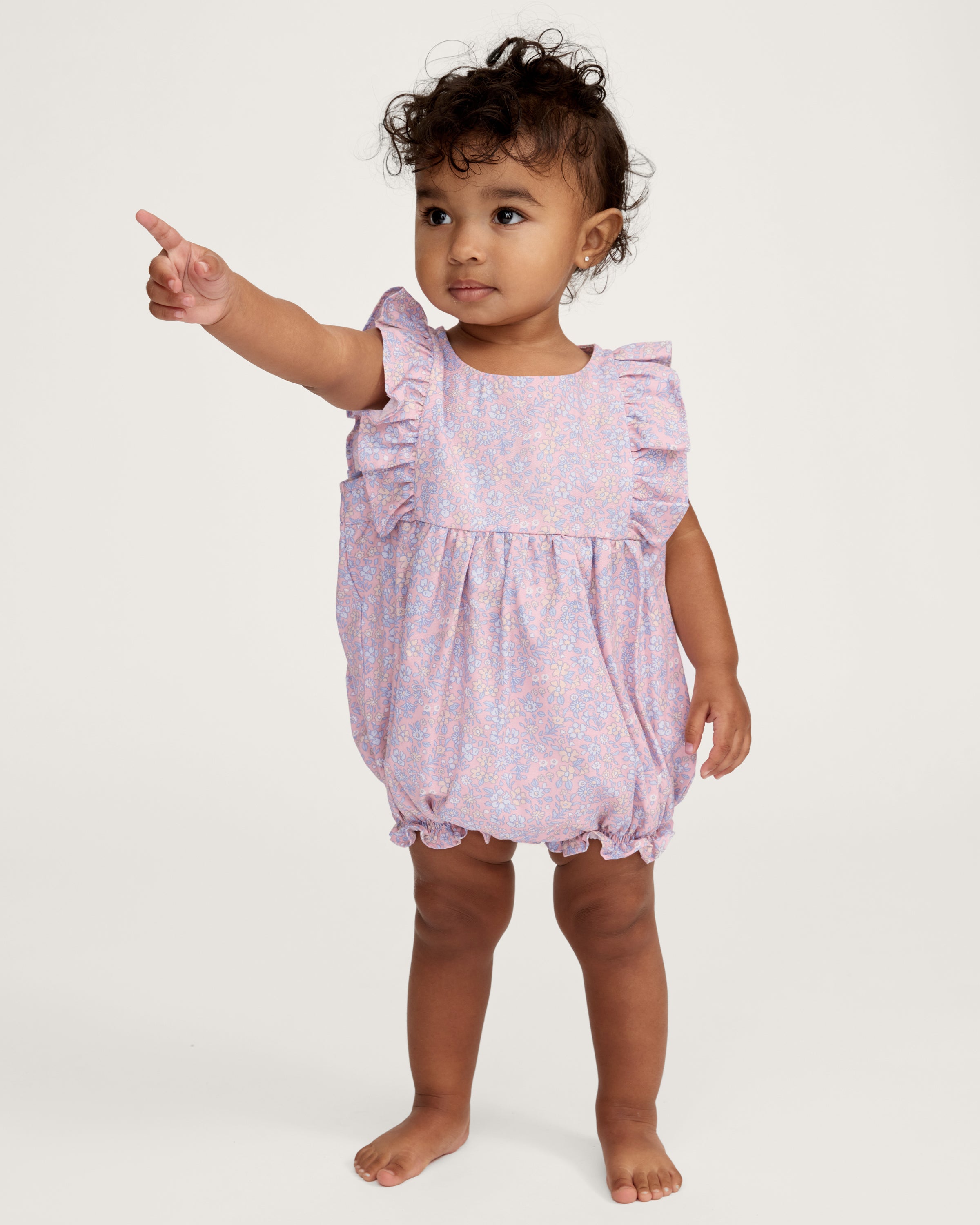 A curly-haired toddler stands barefoot, pointing left and looking that way, wearing the Baby's Twill Ruffled Romper in Fleur Blush against a plain light background.