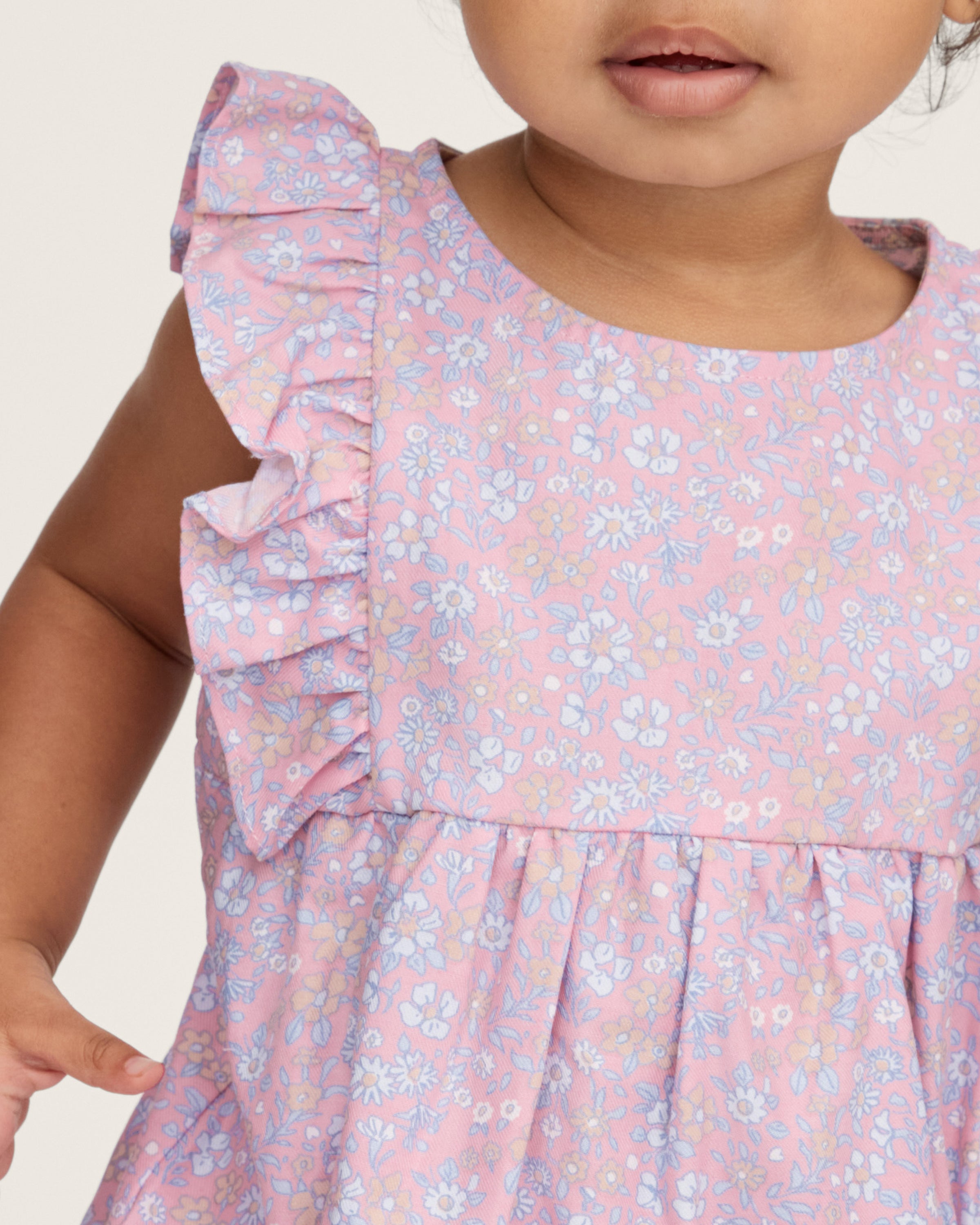 A curly-haired toddler stands barefoot, pointing left and looking that way, wearing the Baby's Twill Ruffled Romper in Fleur Blush against a plain light background.