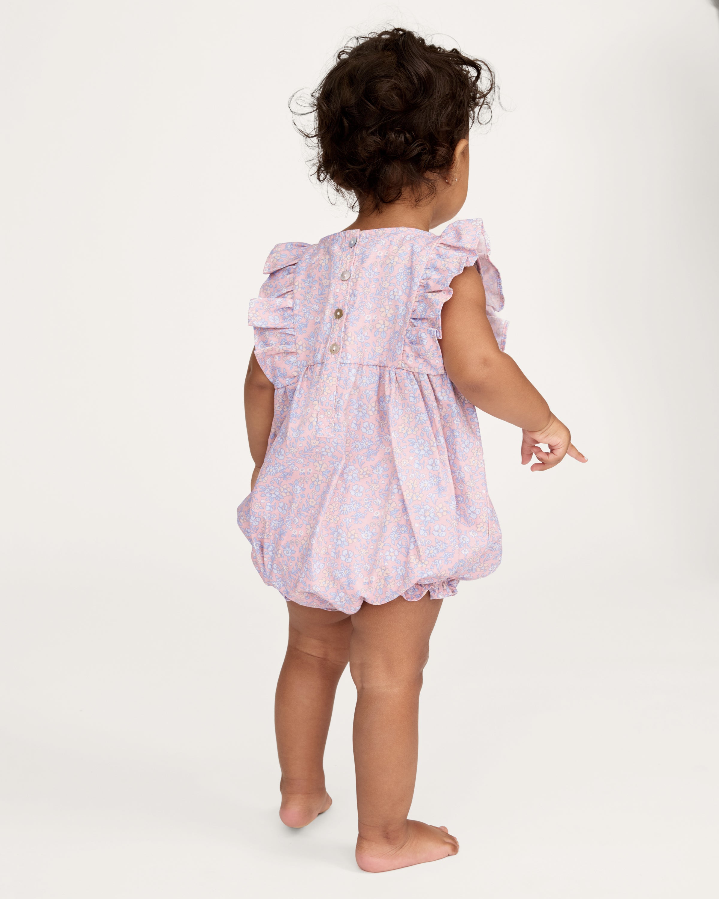 A curly-haired toddler stands barefoot, pointing left and looking that way, wearing the Baby's Twill Ruffled Romper in Fleur Blush against a plain light background.