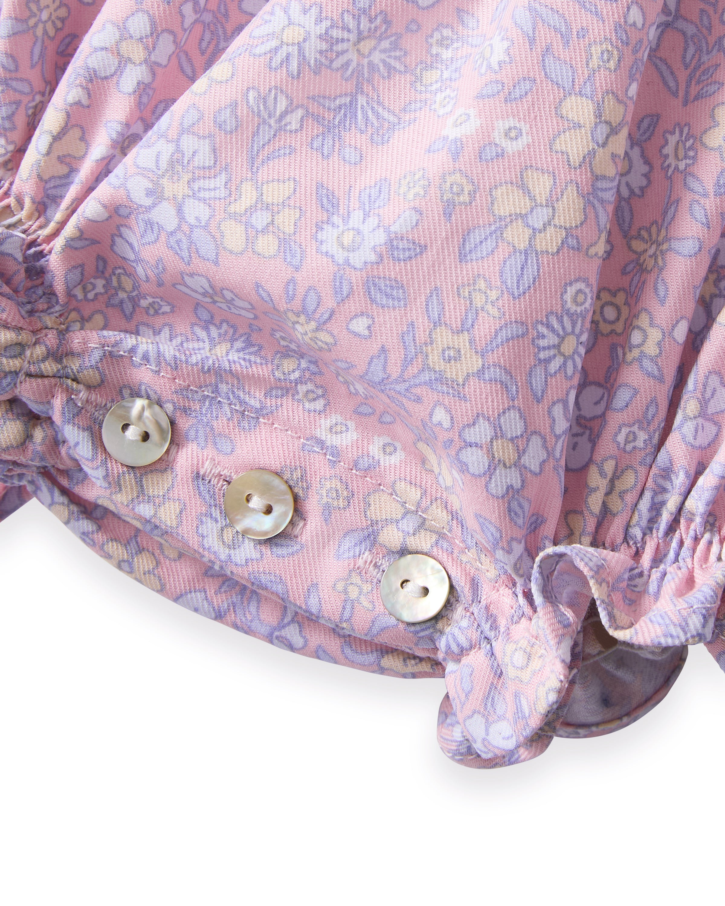 A curly-haired toddler stands barefoot, pointing left and looking that way, wearing the Baby's Twill Ruffled Romper in Fleur Blush against a plain light background.