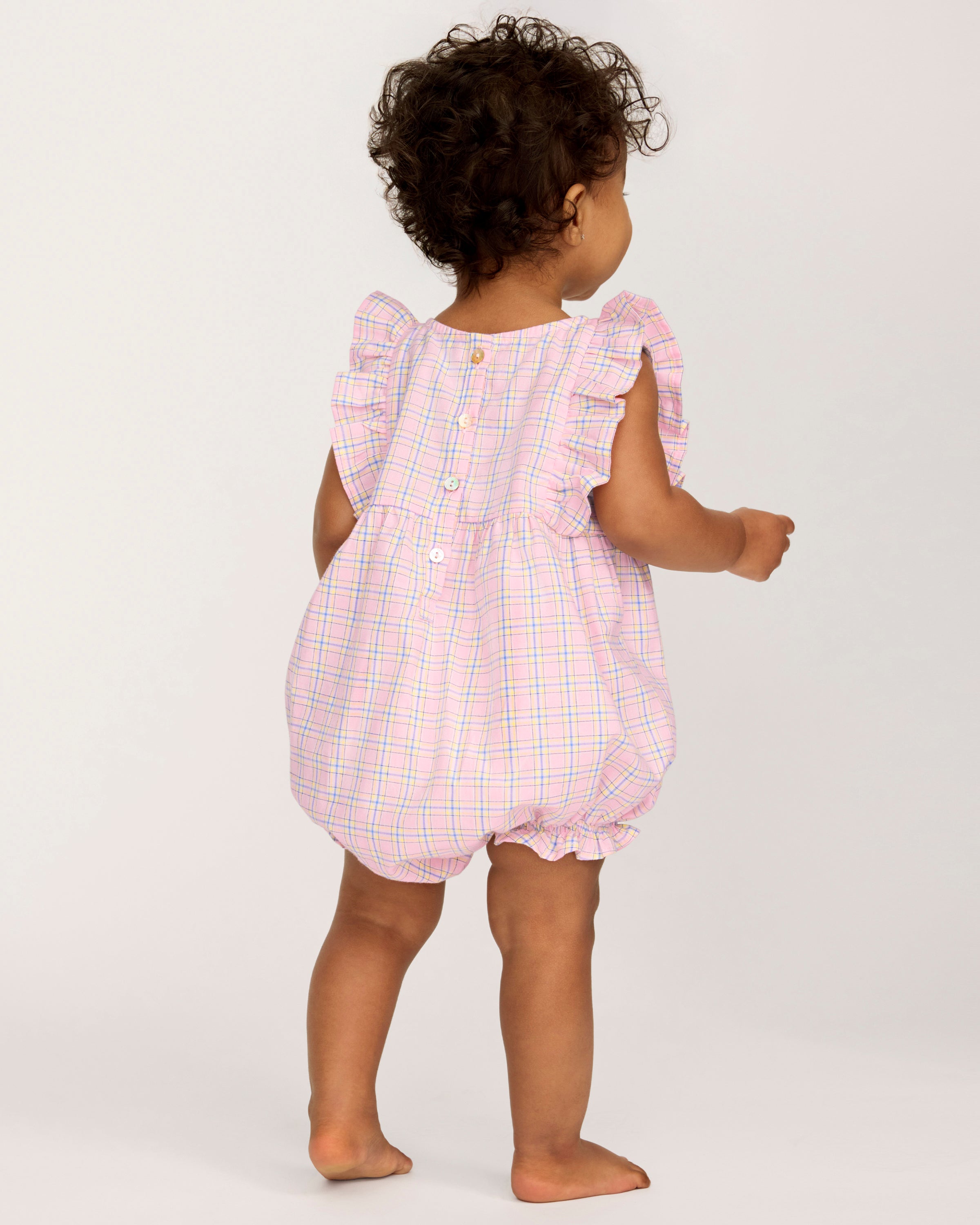 A toddler with curly hair wears the Baby's Twill Ruffled Romper in Peony Plaid, featuring ruffled sleeves, standing barefoot against a plain light background and looking slightly to the side.