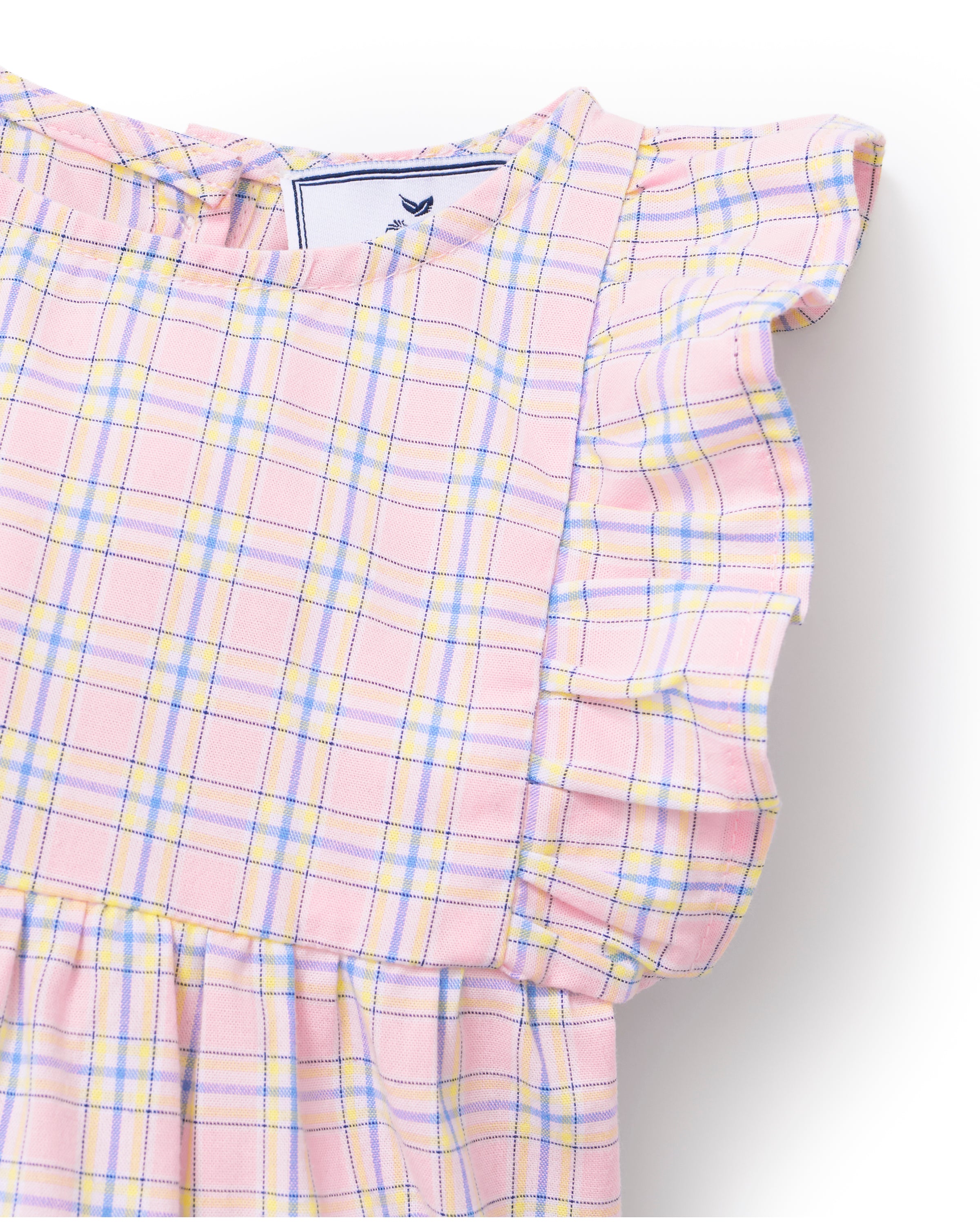 A toddler with curly hair wears the Baby's Twill Ruffled Romper in Peony Plaid, featuring ruffled sleeves, standing barefoot against a plain light background and looking slightly to the side.