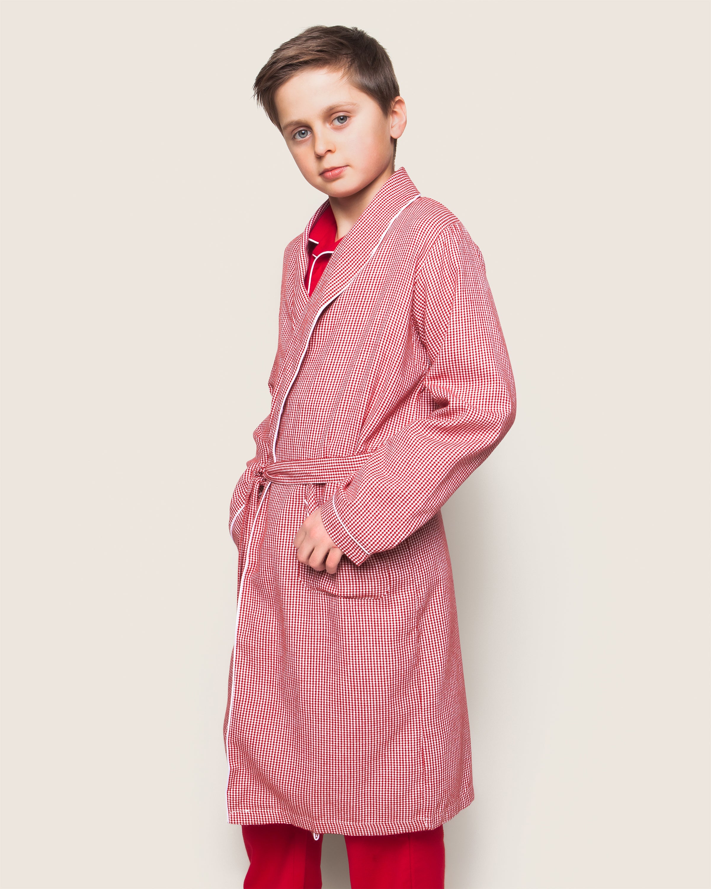 A boy models Petite Plumes Kids Flannel Robe in Red Mini Gingham against a light backdrop. The robe, complete with a belt, highlights his casual vibe as he gazes neutrally at the camera with hands in pockets.