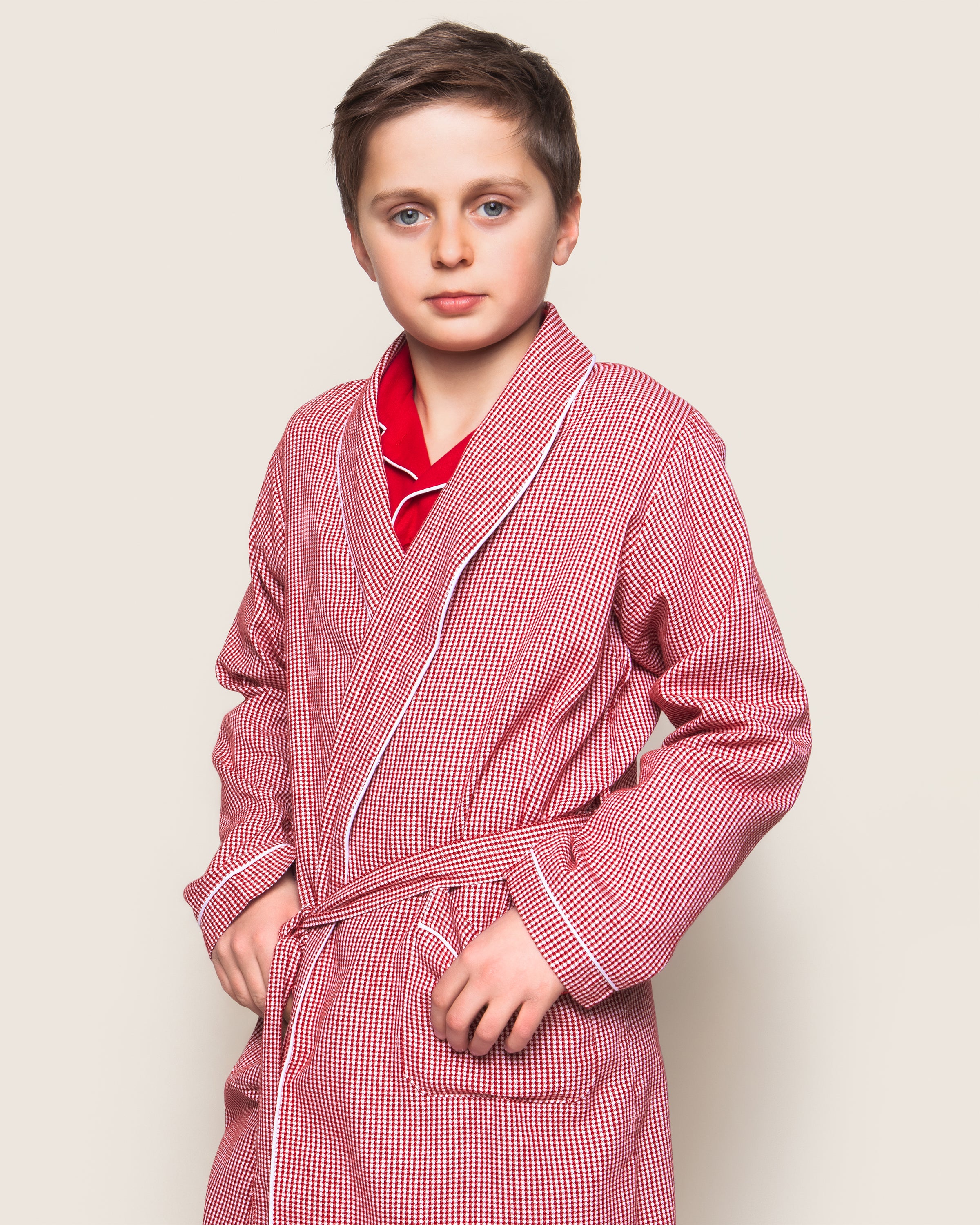 A boy in Petite Plumes Kids Flannel Robe in Red Mini Gingham, made from flame-retardant fabric, stands with hands in pockets. The robe has a waist tie and reveals a red shirt underneath as he looks directly at the camera against a plain background.