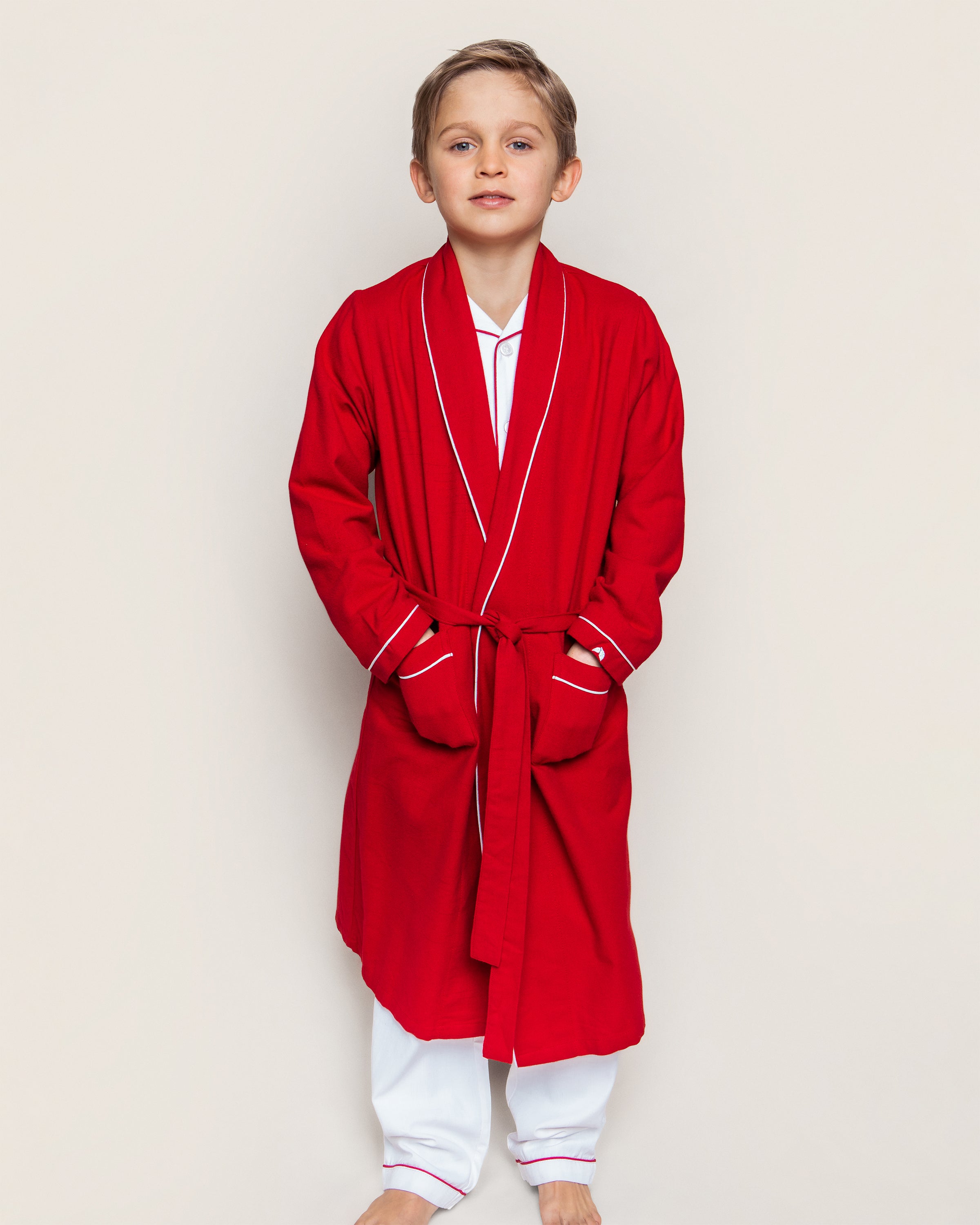 A young boy stands against a plain background, wearing Petite Plumes Kids Flannel Robe in Red with White Piping over pajamas. He has short hair and is barefoot, looking directly at the camera with a neutral expression.