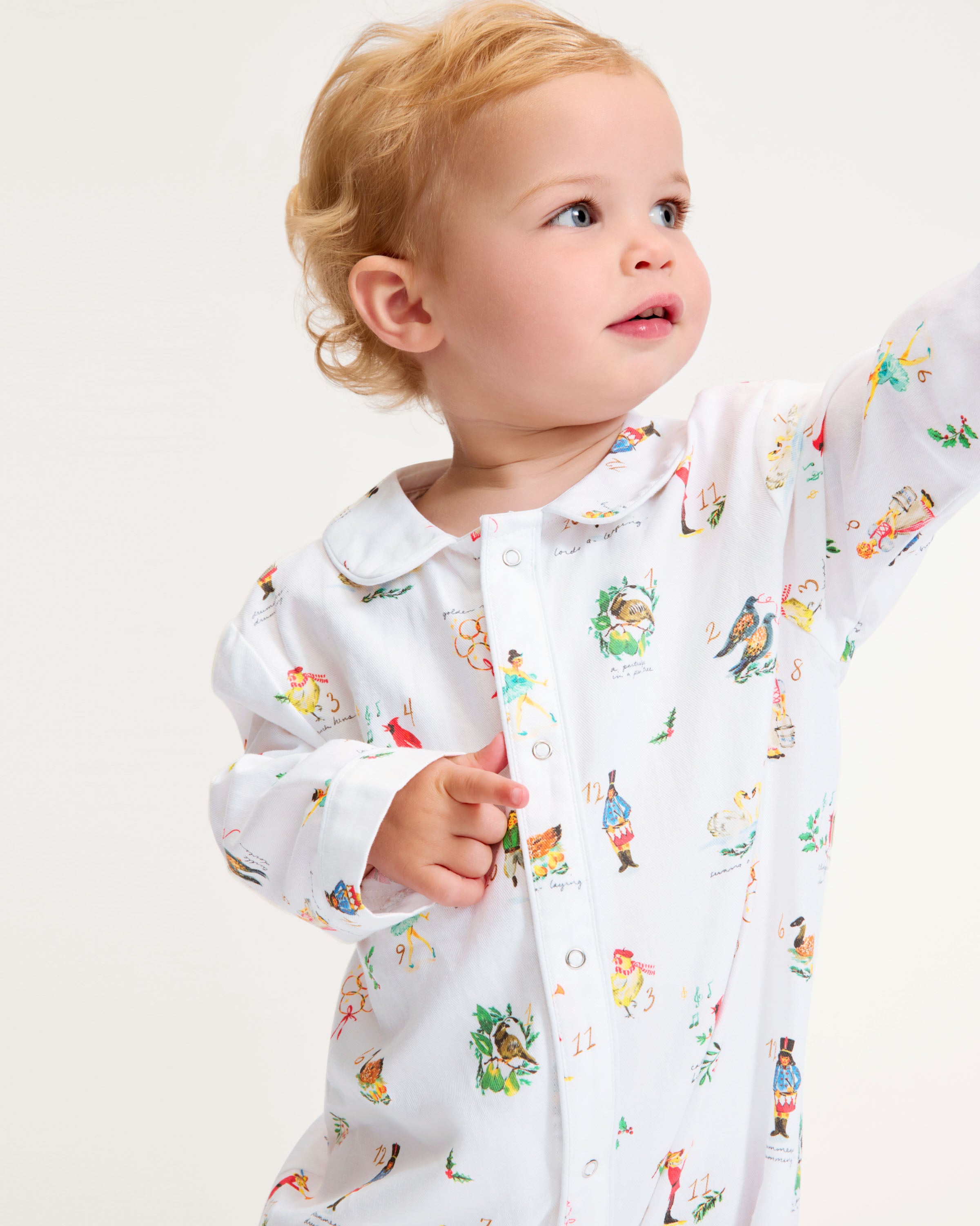 The Baby's Twill Cambridge Romper in 12 Days of Christmas features a white long-sleeve design with a collar, playful festive illustrations, and convenient snap buttons down the front and legs.