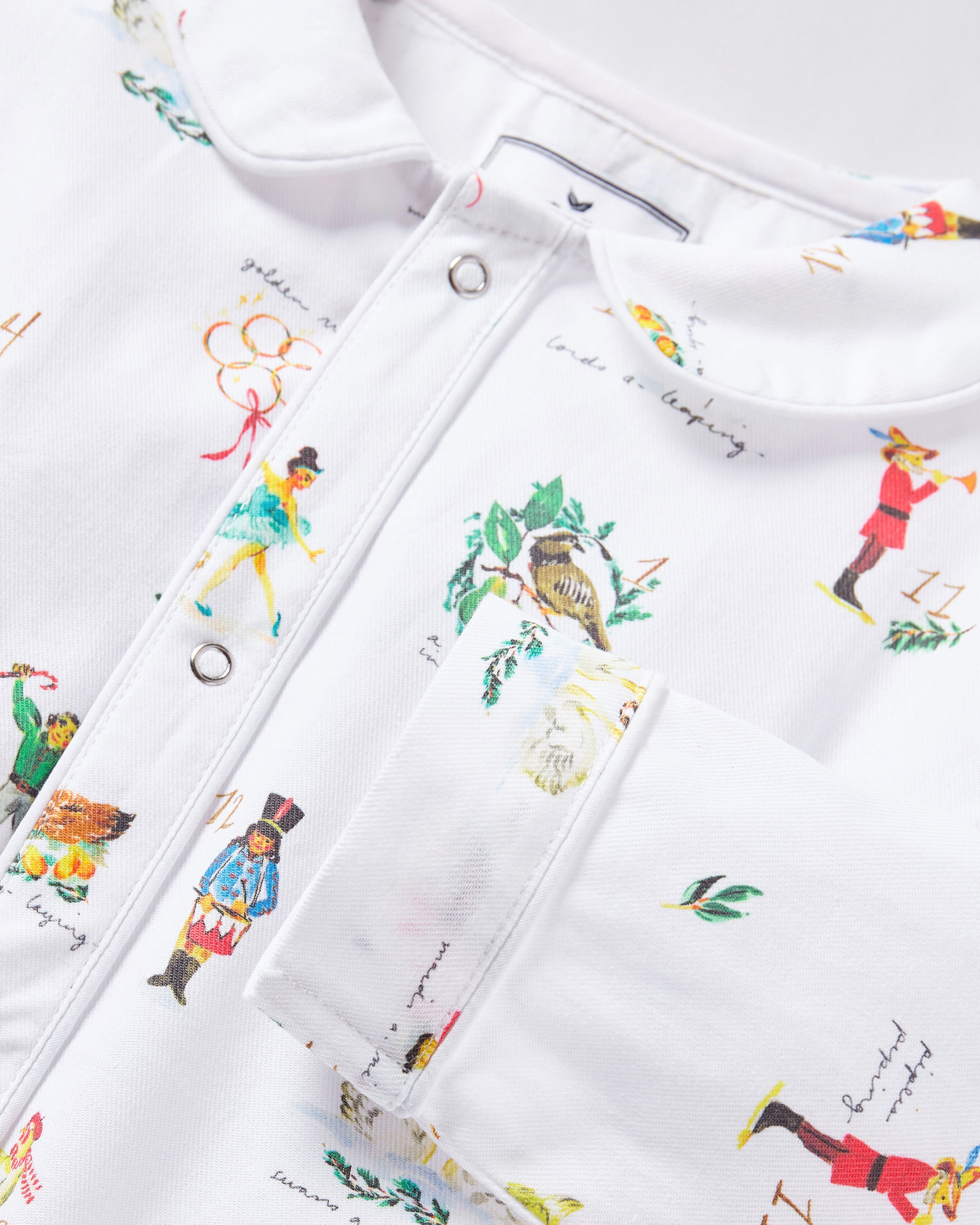 The Baby's Twill Cambridge Romper in 12 Days of Christmas features a white long-sleeve design with a collar, playful festive illustrations, and convenient snap buttons down the front and legs.