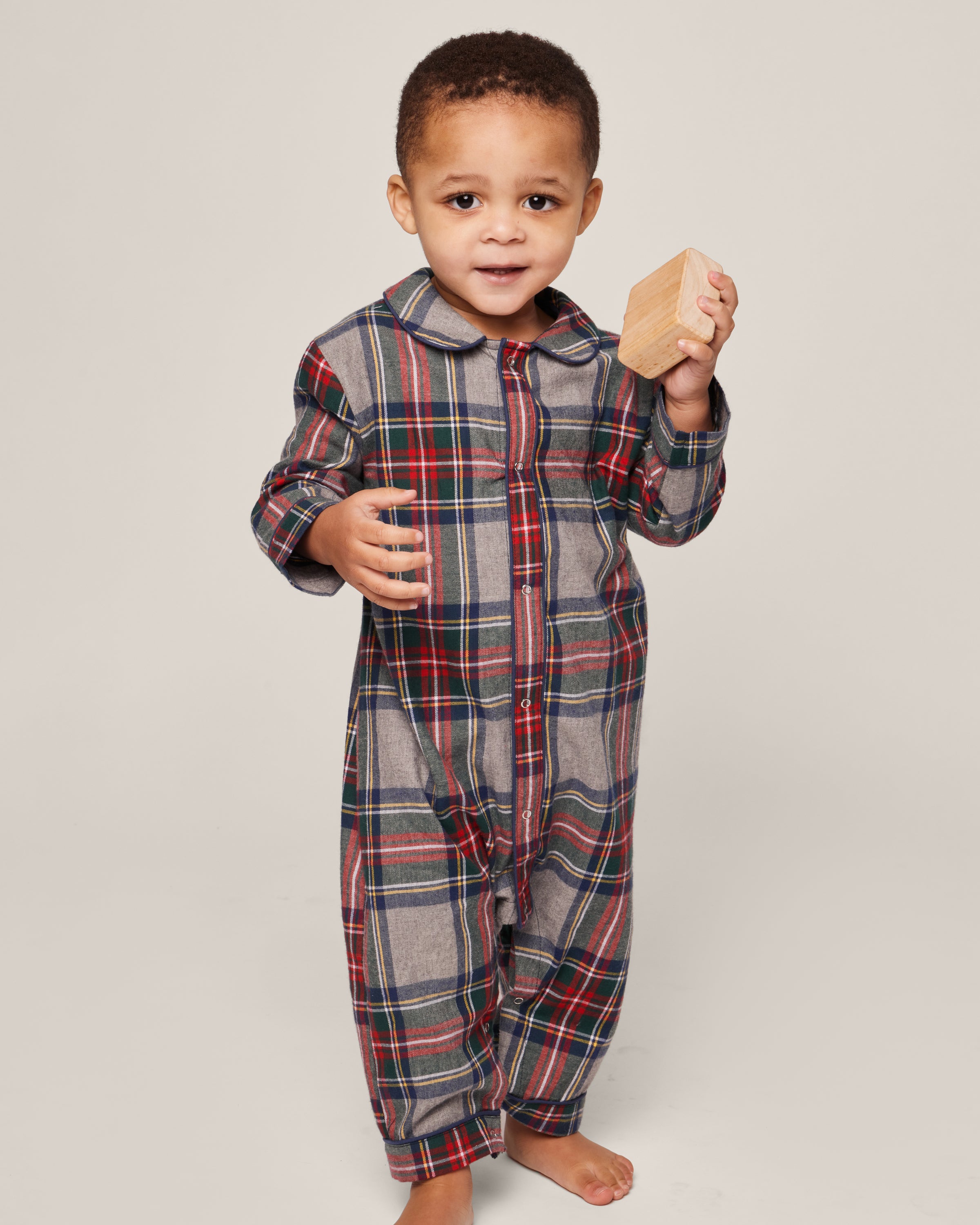 A small child in the Petite Plume Babys Brushed Cotton Cambridge Romper, featuring Westminster Tartan with red, green, and blue stripes, stands barefoot holding a wooden block against a plain light-colored background.