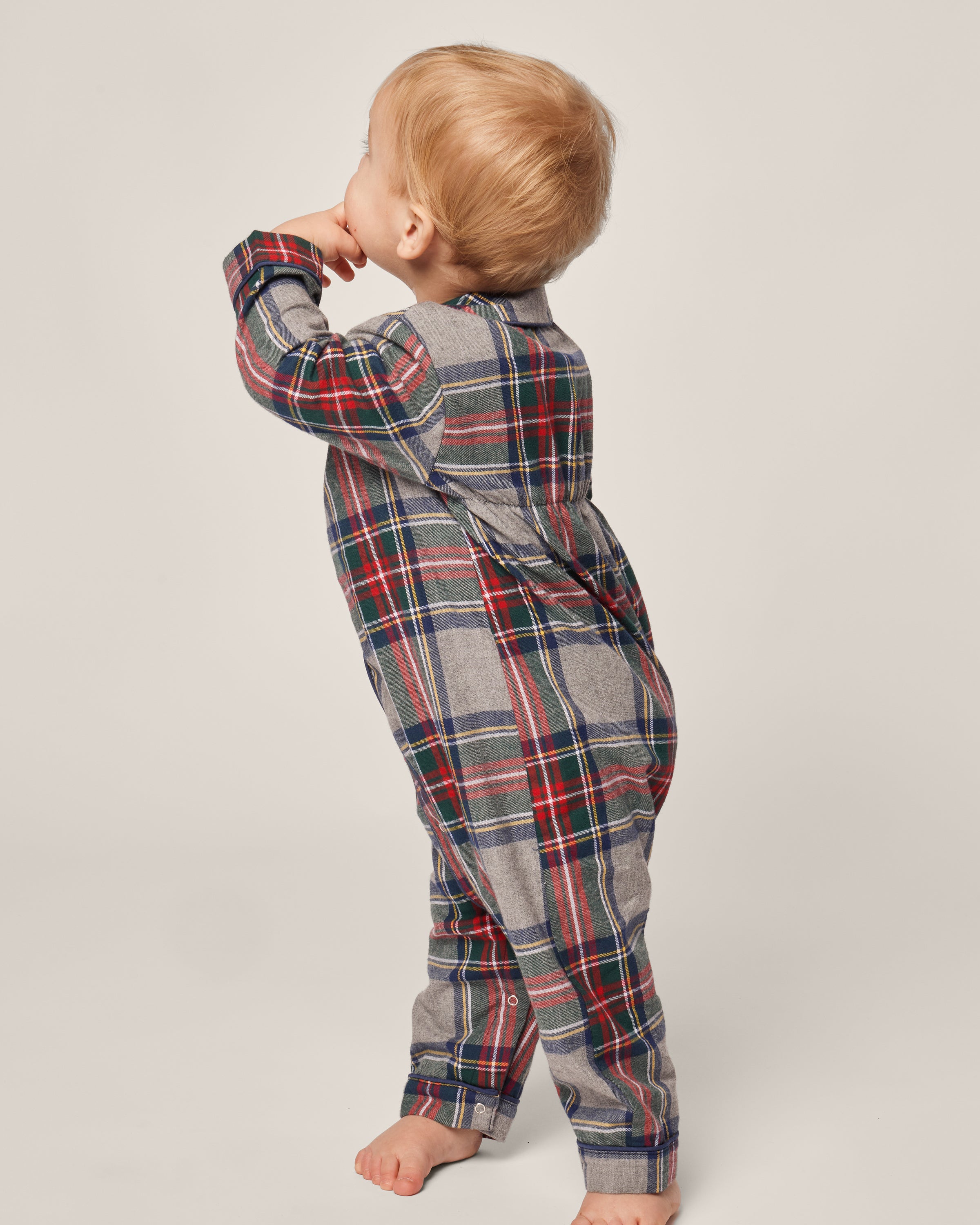 A toddler wearing a Babys Brushed Cotton Cambridge Romper in Westminster Tartan by Petite Plume stands barefoot on a light background, facing away with their right thumb in their mouth and left hand slightly raised. The romper features a traditional blue, red, and green plaid motif.