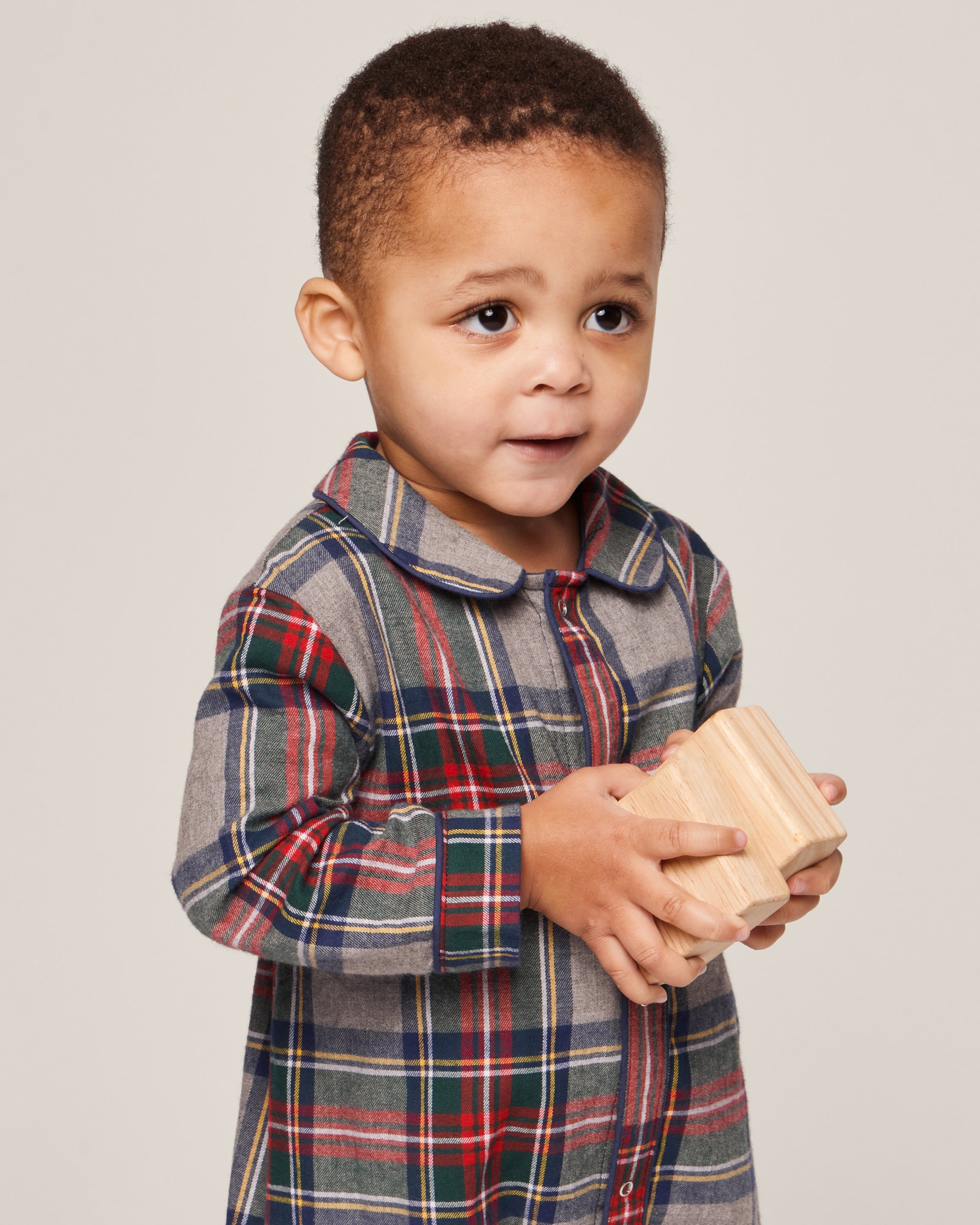 In a Babys Brushed Cotton Cambridge Romper in Westminster Tartan by Petite Plume, a young child thoughtfully holds a wooden block against a plain background.