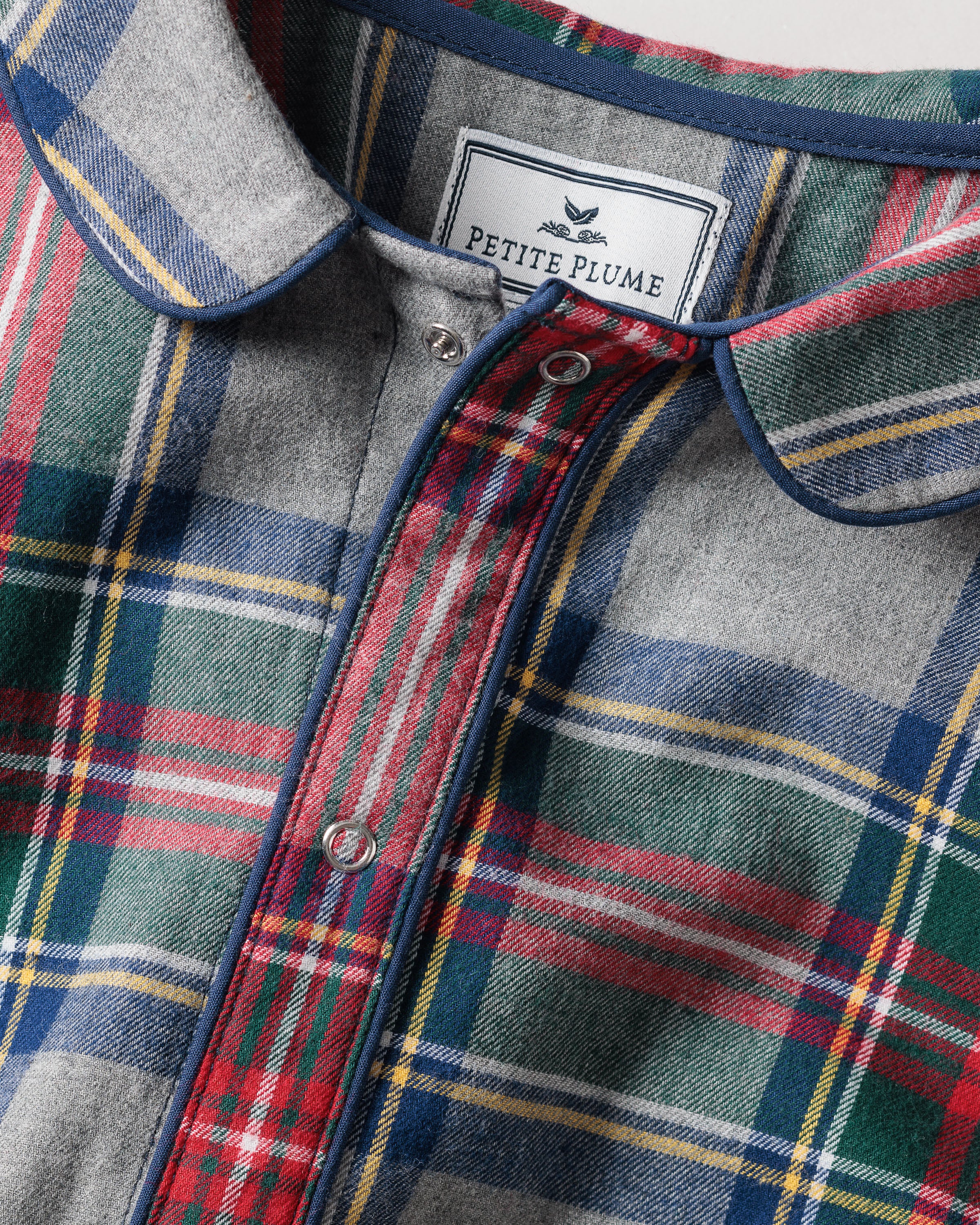 Close-up of Petite Plumes Babys Brushed Cotton Cambridge Romper in Westminster Tartan featuring a button-up front in red, green, blue, and yellow on a gray background, inspired by Scottish tradition. Perfect for childrens sleepwear with its cozy design and small bird illustration on the label.