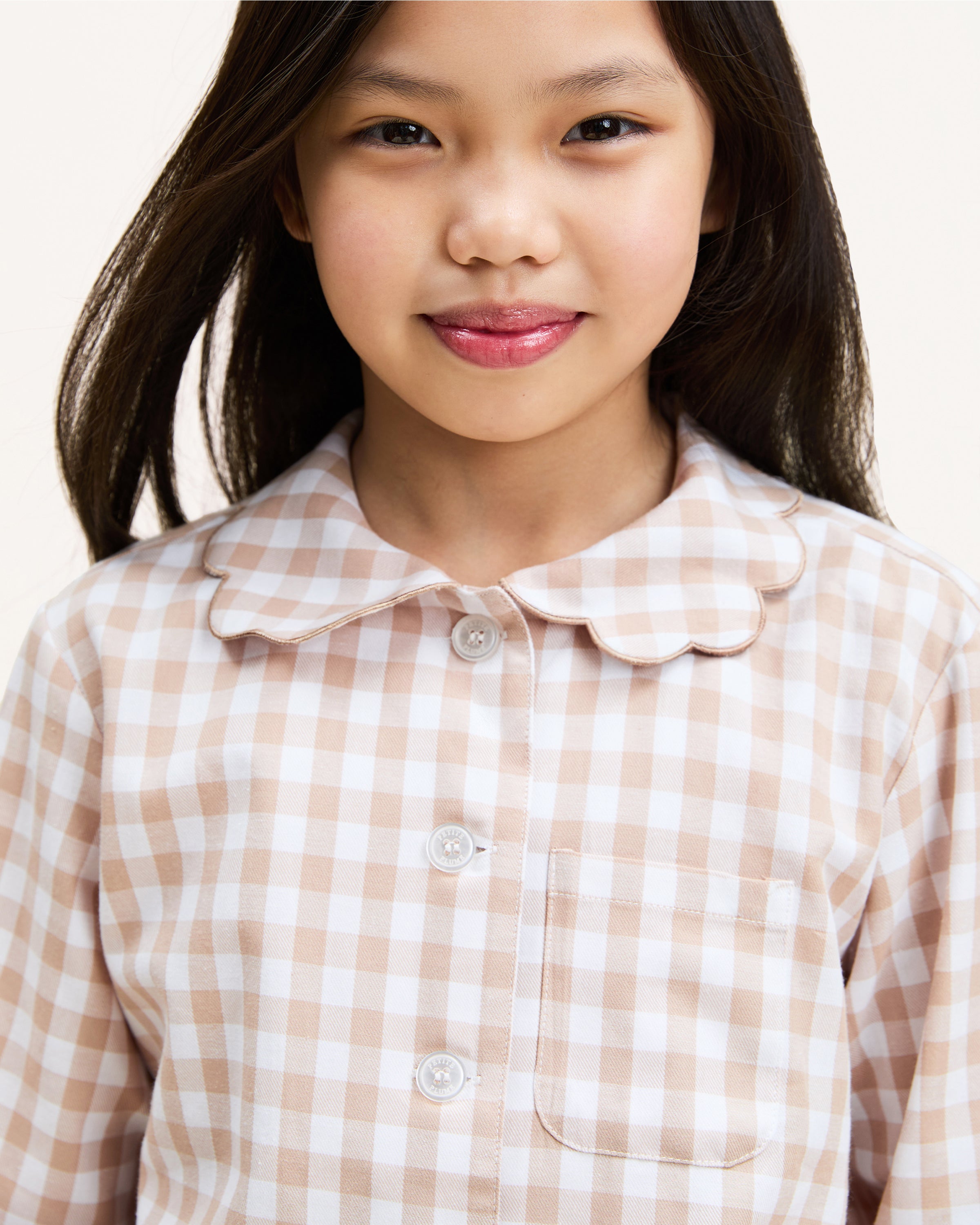 Kid's Twill Colette Pajama Set in Camel Gingham, featuring a beige and white gingham pattern with a scalloped collar, long sleeves, front pocket, and matching elastic-waist pants, is displayed neatly on a white background.