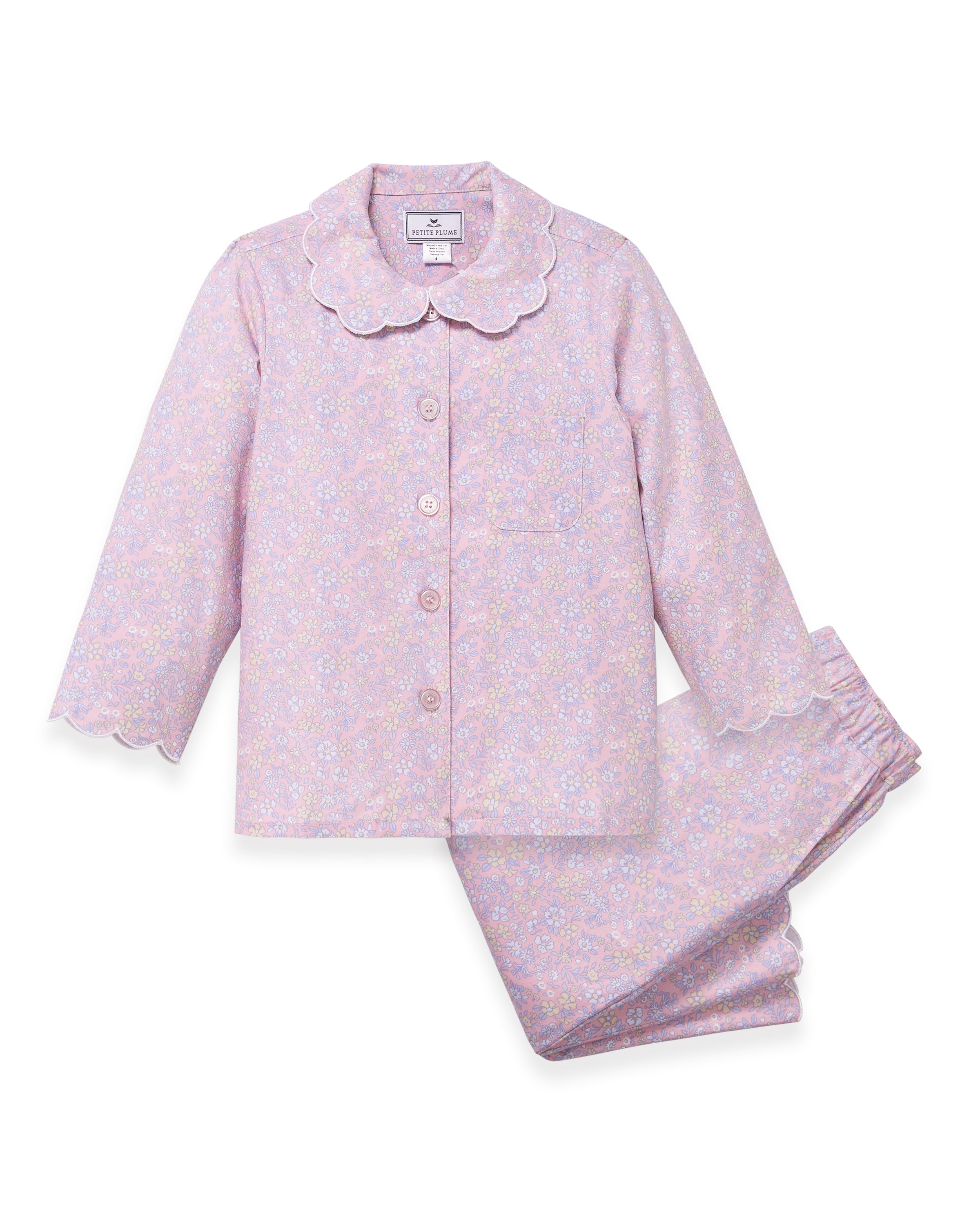 A young girl with wavy hair stands barefoot in the Kid's Twill Colette Pajama Set in Fleur Blush, featuring light pink floral fabric with a scalloped collar and pants, posing against a plain white background.