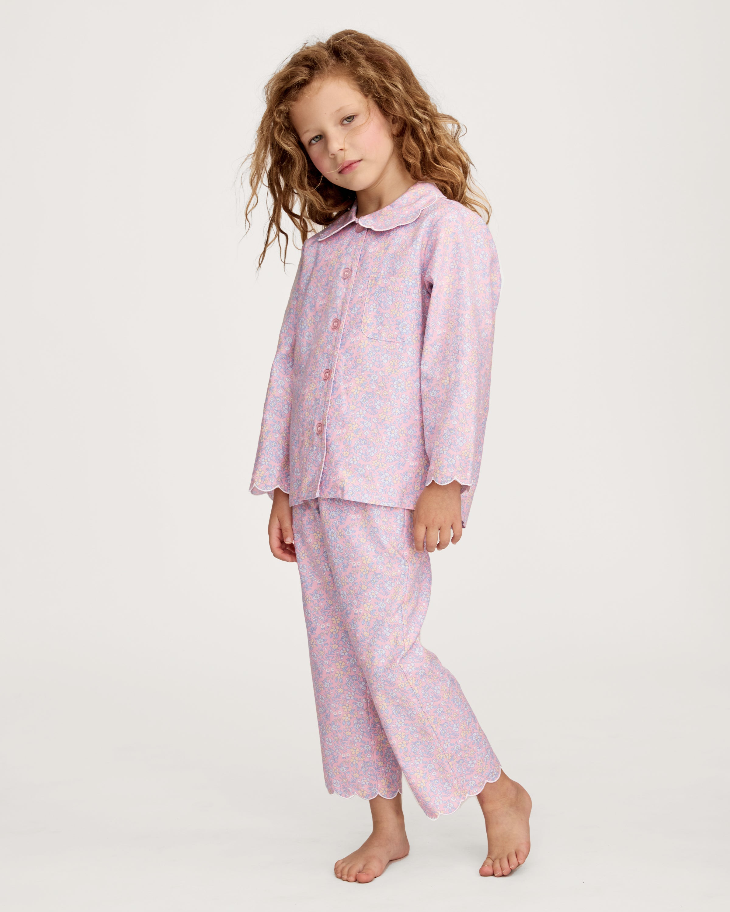 A young girl with wavy hair stands barefoot in the Kid's Twill Colette Pajama Set in Fleur Blush, featuring light pink floral fabric with a scalloped collar and pants, posing against a plain white background.