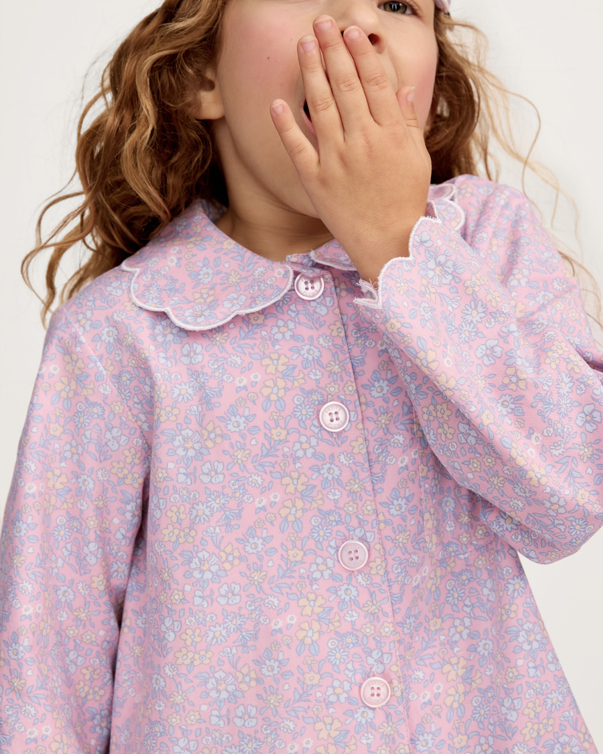 A young girl with wavy hair stands barefoot in the Kid's Twill Colette Pajama Set in Fleur Blush, featuring light pink floral fabric with a scalloped collar and pants, posing against a plain white background.