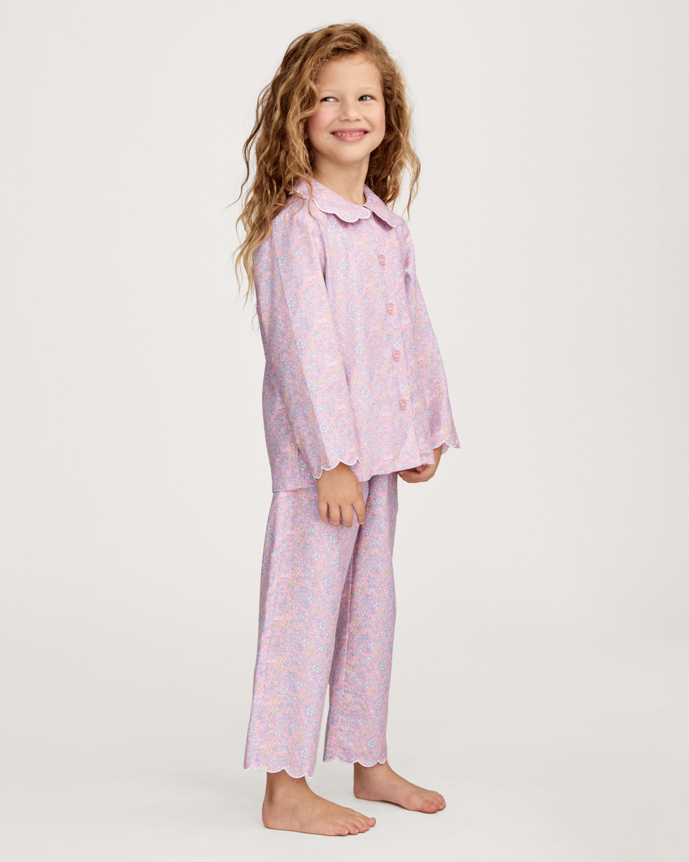 A young girl with wavy hair stands barefoot in the Kid's Twill Colette Pajama Set in Fleur Blush, featuring light pink floral fabric with a scalloped collar and pants, posing against a plain white background.