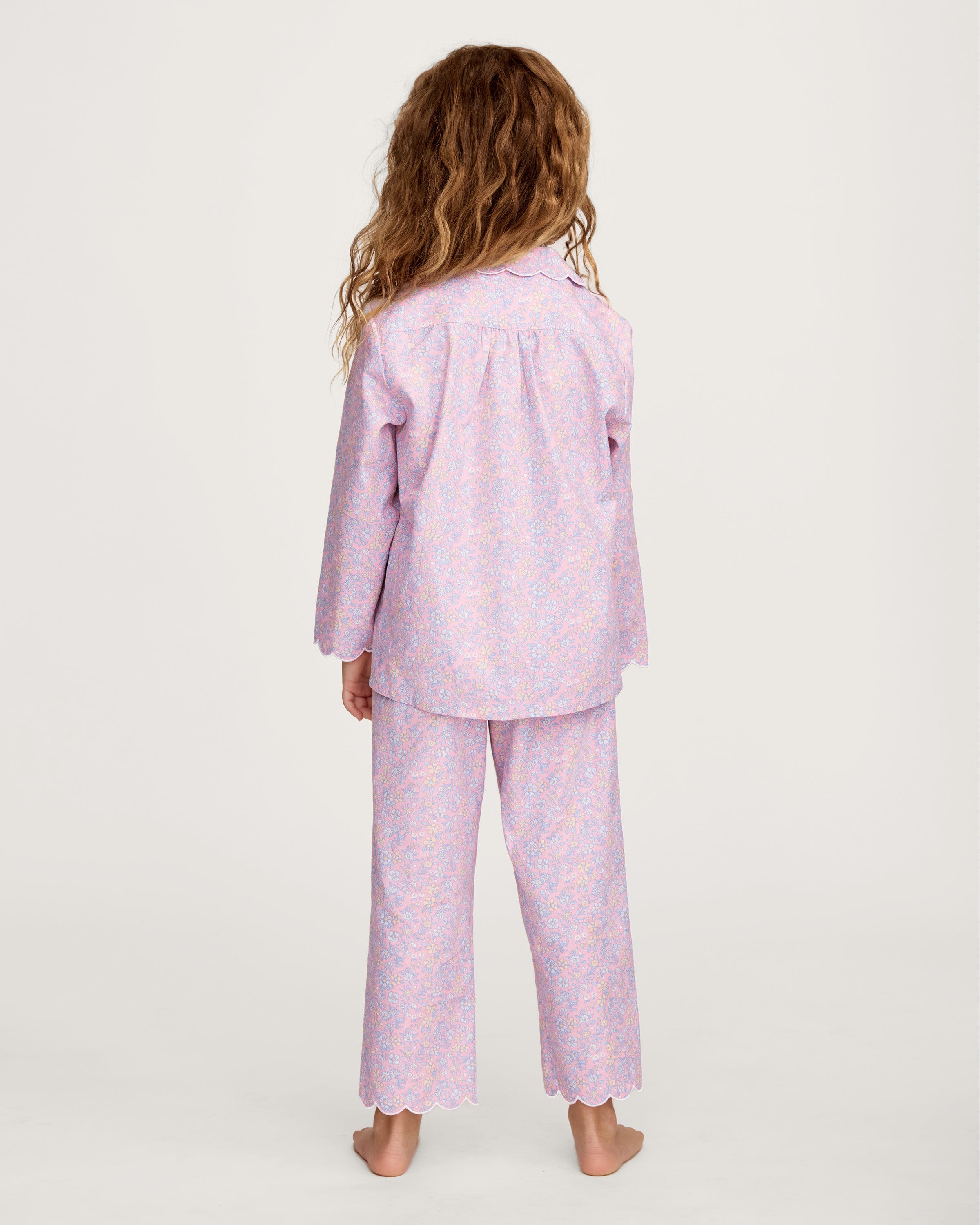 A young girl with wavy hair stands barefoot in the Kid's Twill Colette Pajama Set in Fleur Blush, featuring light pink floral fabric with a scalloped collar and pants, posing against a plain white background.