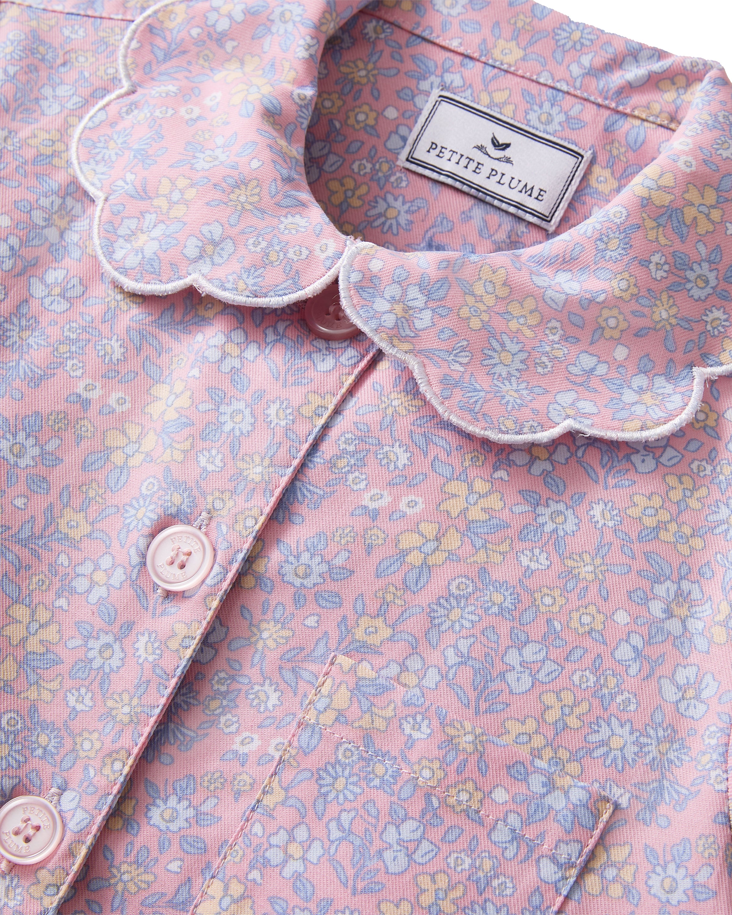 A young girl with wavy hair stands barefoot in the Kid's Twill Colette Pajama Set in Fleur Blush, featuring light pink floral fabric with a scalloped collar and pants, posing against a plain white background.
