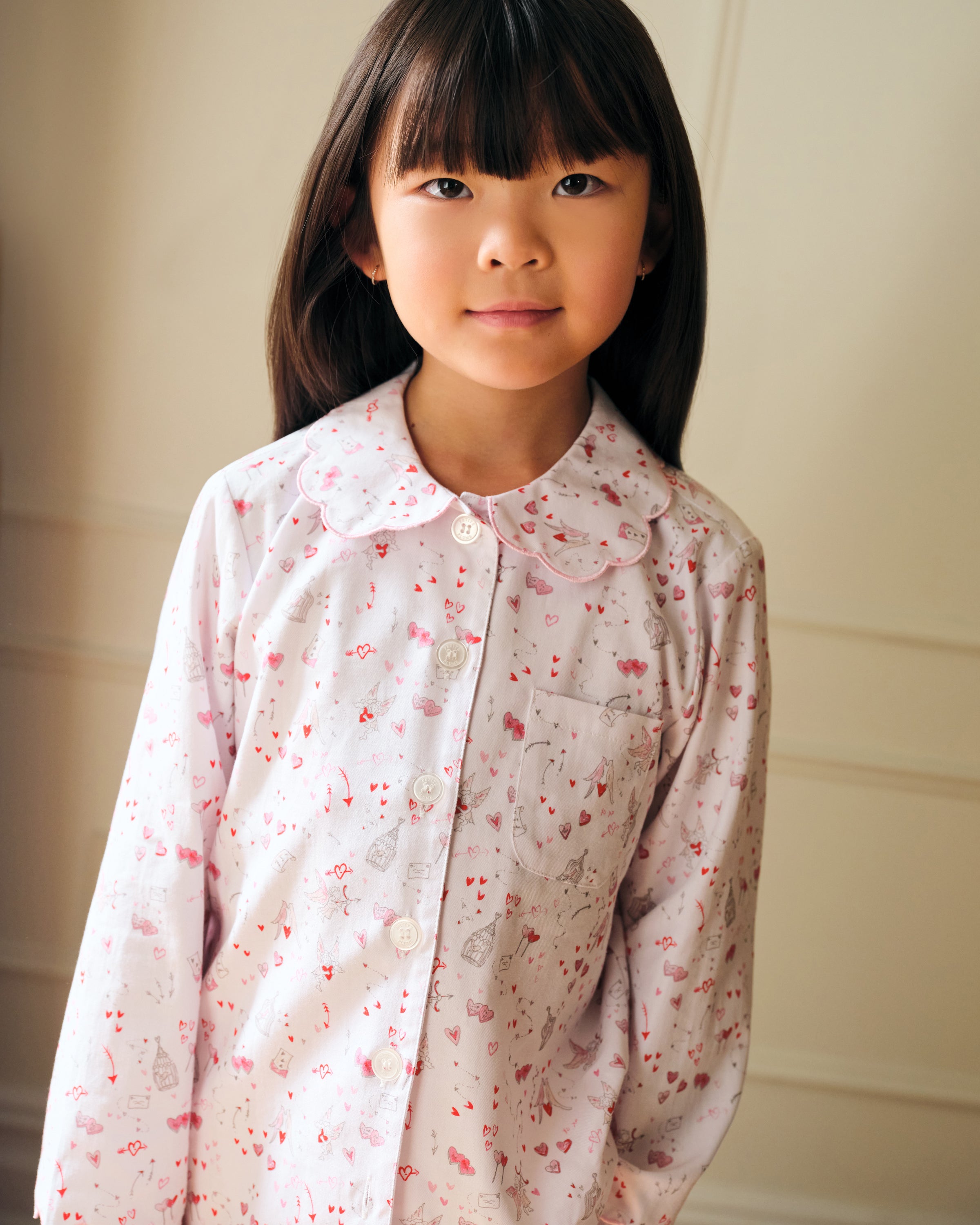The Kid's Twill Colette Pajama Set in Love Song features white pajamas with a scalloped collar and cuffs, adorned with pink and red heart and dot patterns. The set includes a long-sleeved button-up top and matching pants.