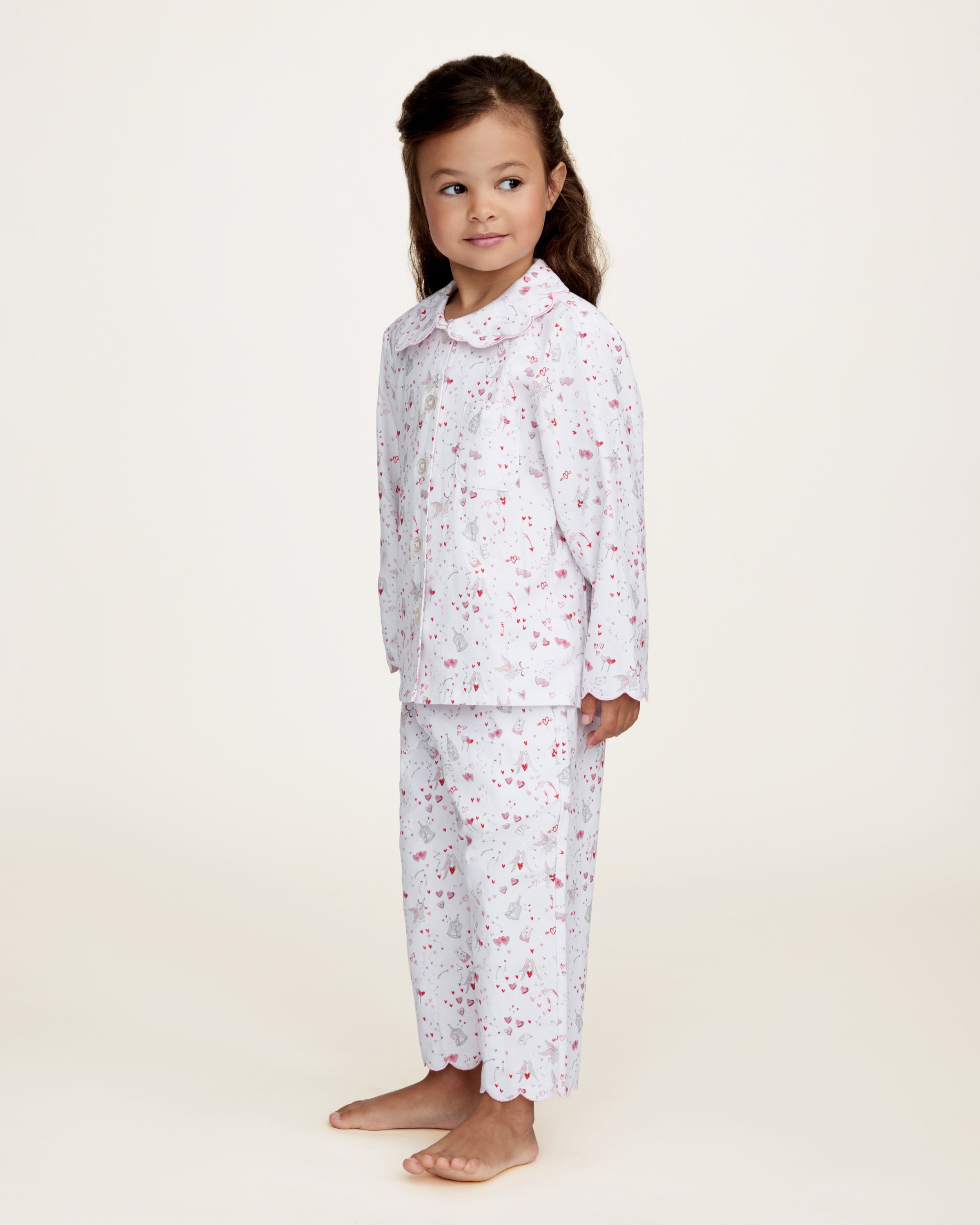 The Kid's Twill Colette Pajama Set in Love Song features white pajamas with a scalloped collar and cuffs, adorned with pink and red heart and dot patterns. The set includes a long-sleeved button-up top and matching pants.