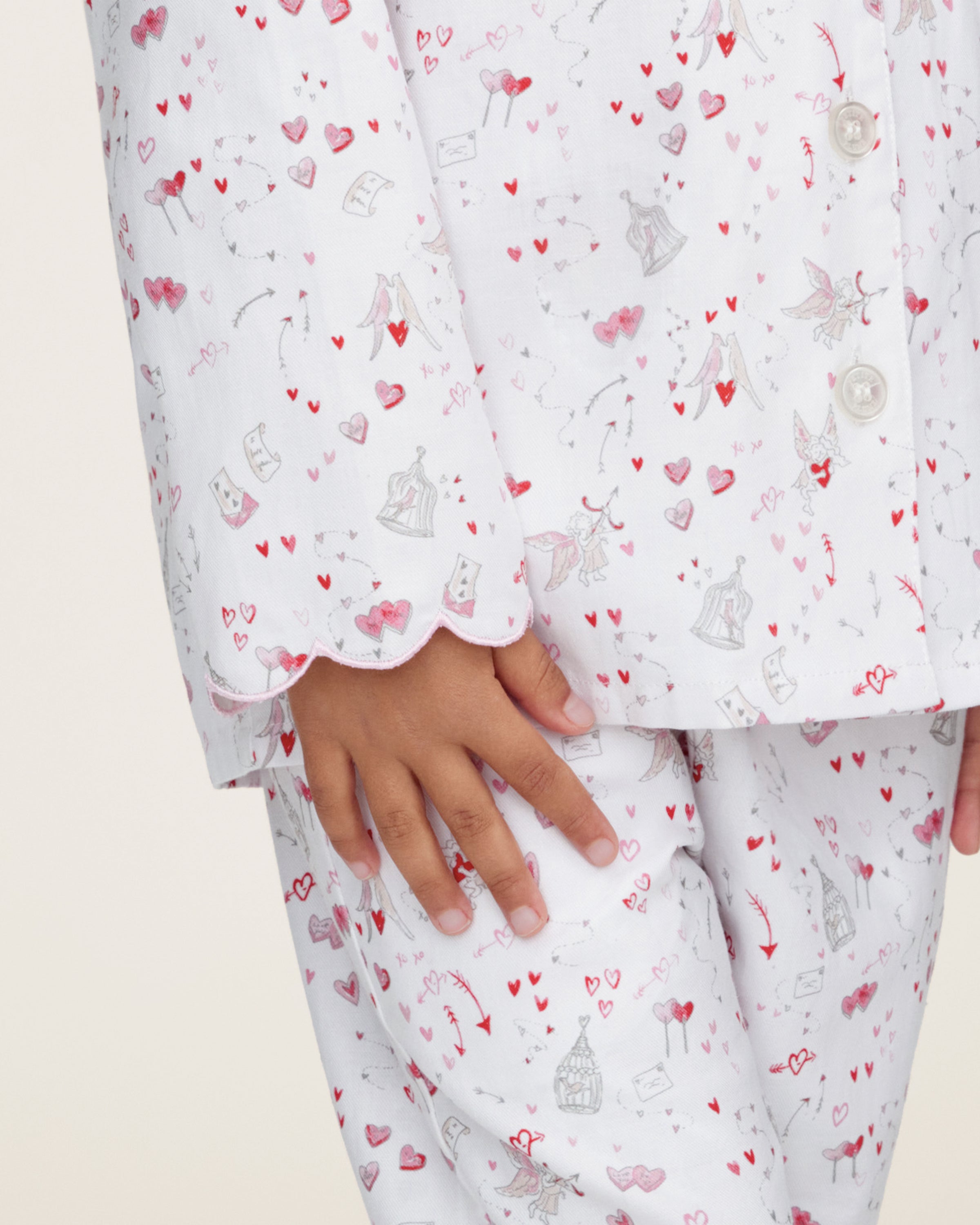 The Kid's Twill Colette Pajama Set in Love Song features white pajamas with a scalloped collar and cuffs, adorned with pink and red heart and dot patterns. The set includes a long-sleeved button-up top and matching pants.