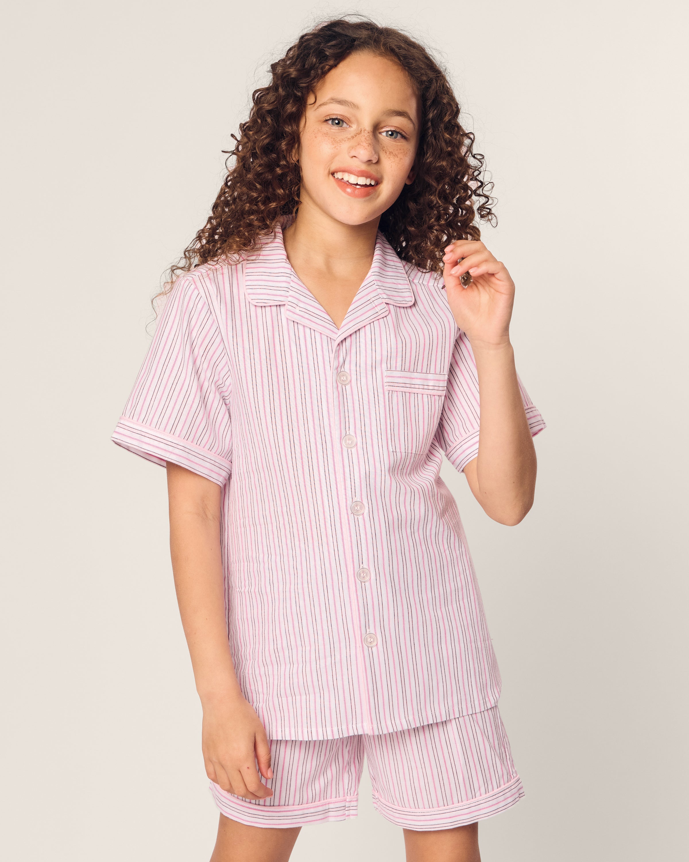 A young girl with curly hair smiles, wearing the Kids Twill Short Set in Blush Boulevard by Petite Plume. The luxury cotton sleepwear features a pink and white striped button-up top with short sleeves and matching shorts, set against a plain beige background, exuding comfort and style.