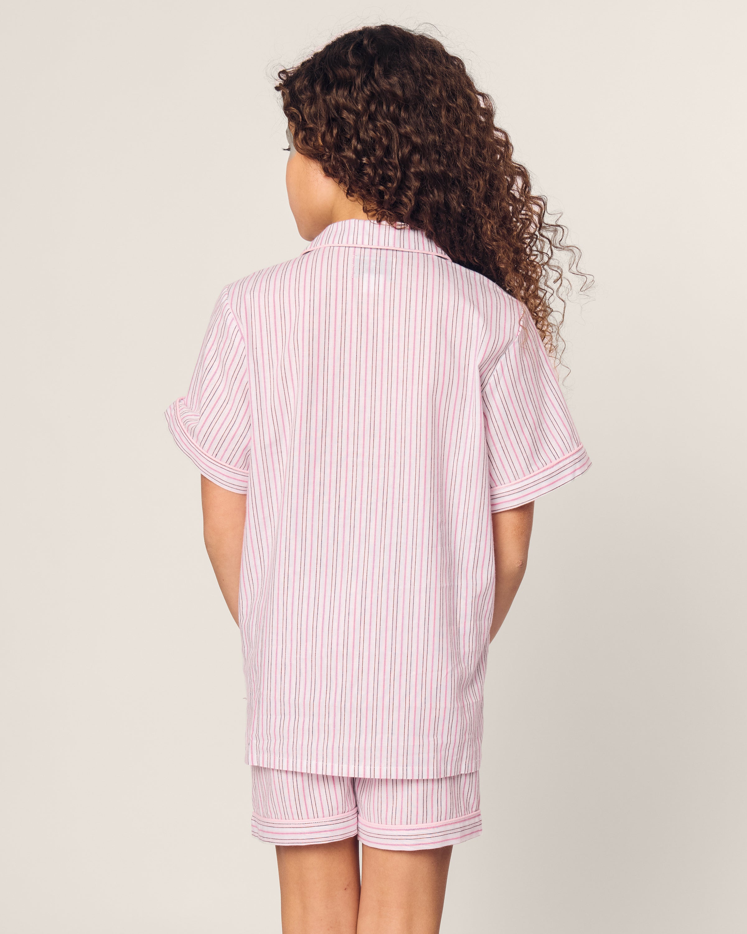 A child with curly brown hair, facing away, wears Petite Plumes Kids Twill Short Set in Blush Boulevard, a luxury cotton sleepwear set featuring pink and white stripes on a short-sleeved shirt and shorts, against a plain light background.