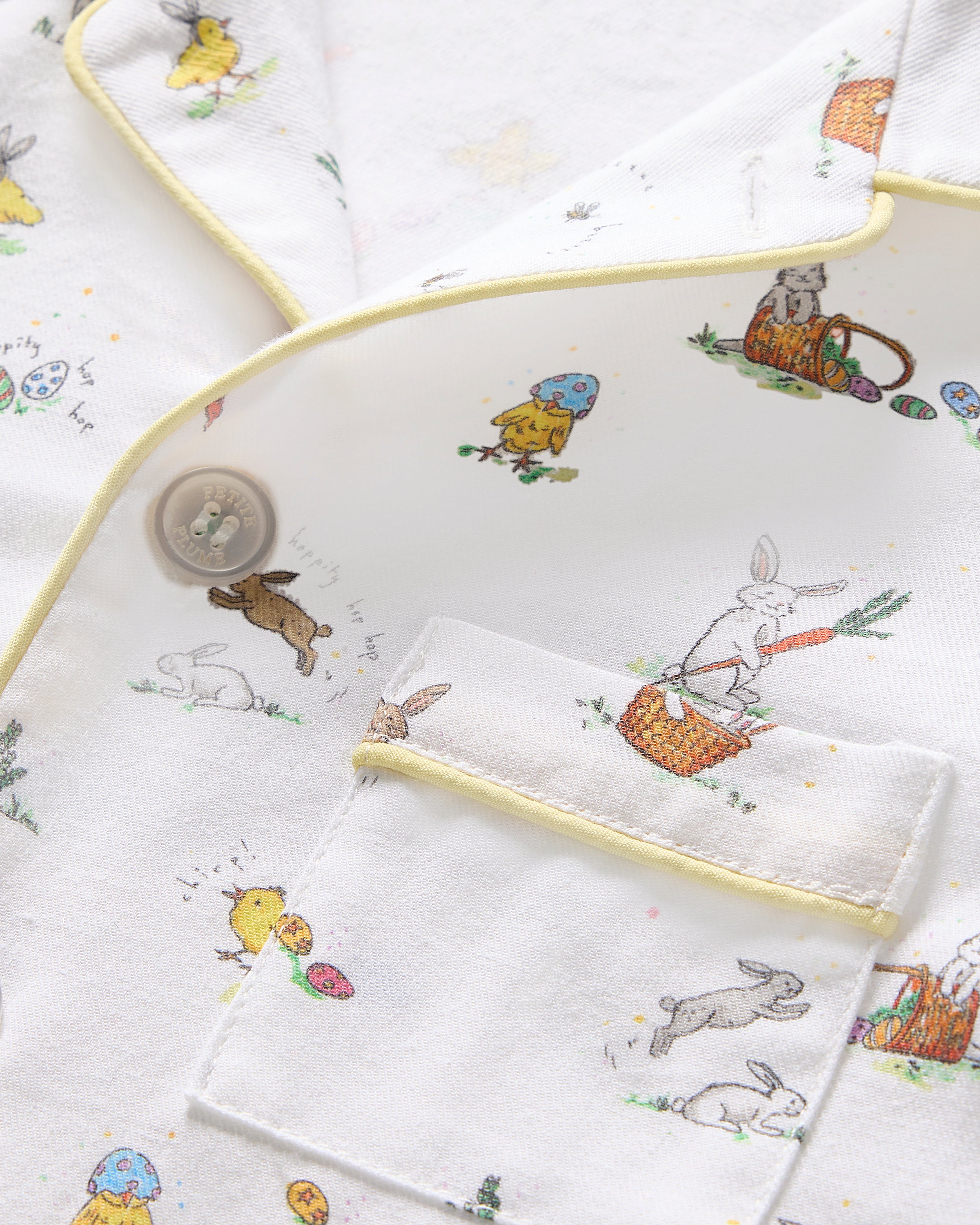 Two smiling children in luxurious Petite Plume sleepwear sit on the floor. They sport matching Kids Twill Short Sets in an Easter Frolic print. One child cuddles a stuffed bunny, embodying comfort and joy against a plain backdrop.