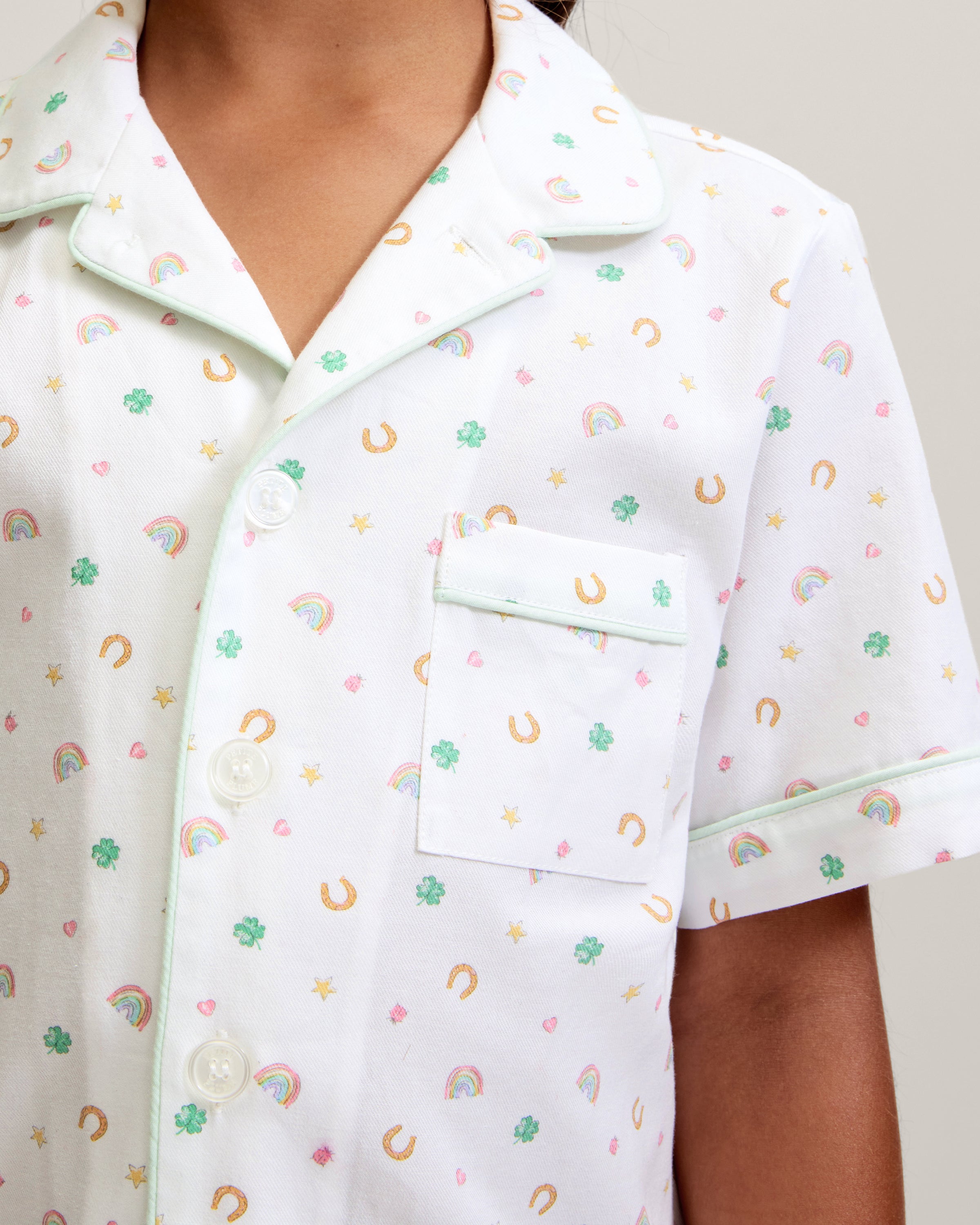 A close-up of a child in the Kid's Twill Pajama Short Set in Good Luck Charms, featuring white fabric with light green trim and pastel rainbows, horseshoes, four-leaf clovers, and stars. Only the upper torso is shown.