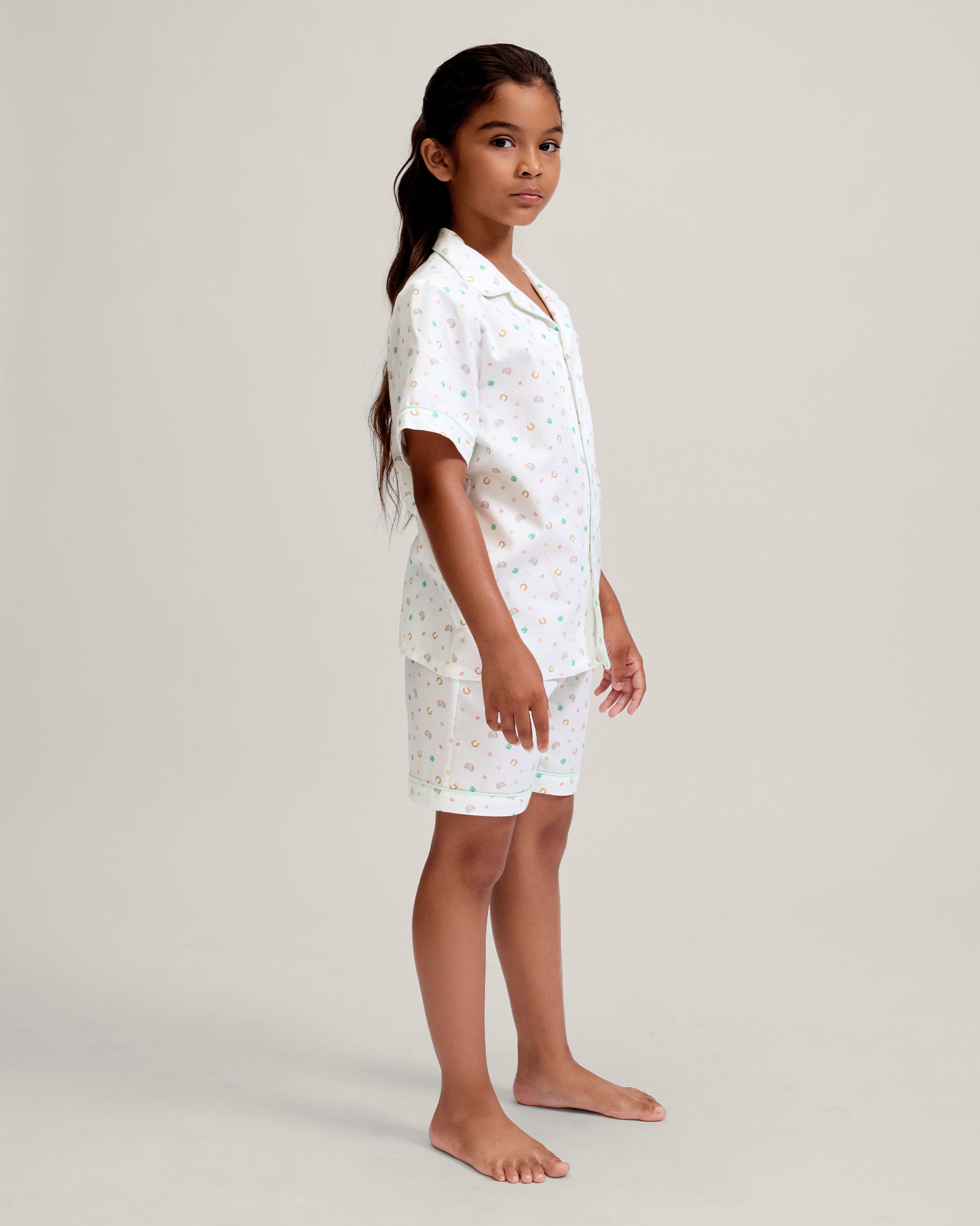 A close-up of a child in the Kid's Twill Pajama Short Set in Good Luck Charms, featuring white fabric with light green trim and pastel rainbows, horseshoes, four-leaf clovers, and stars. Only the upper torso is shown.