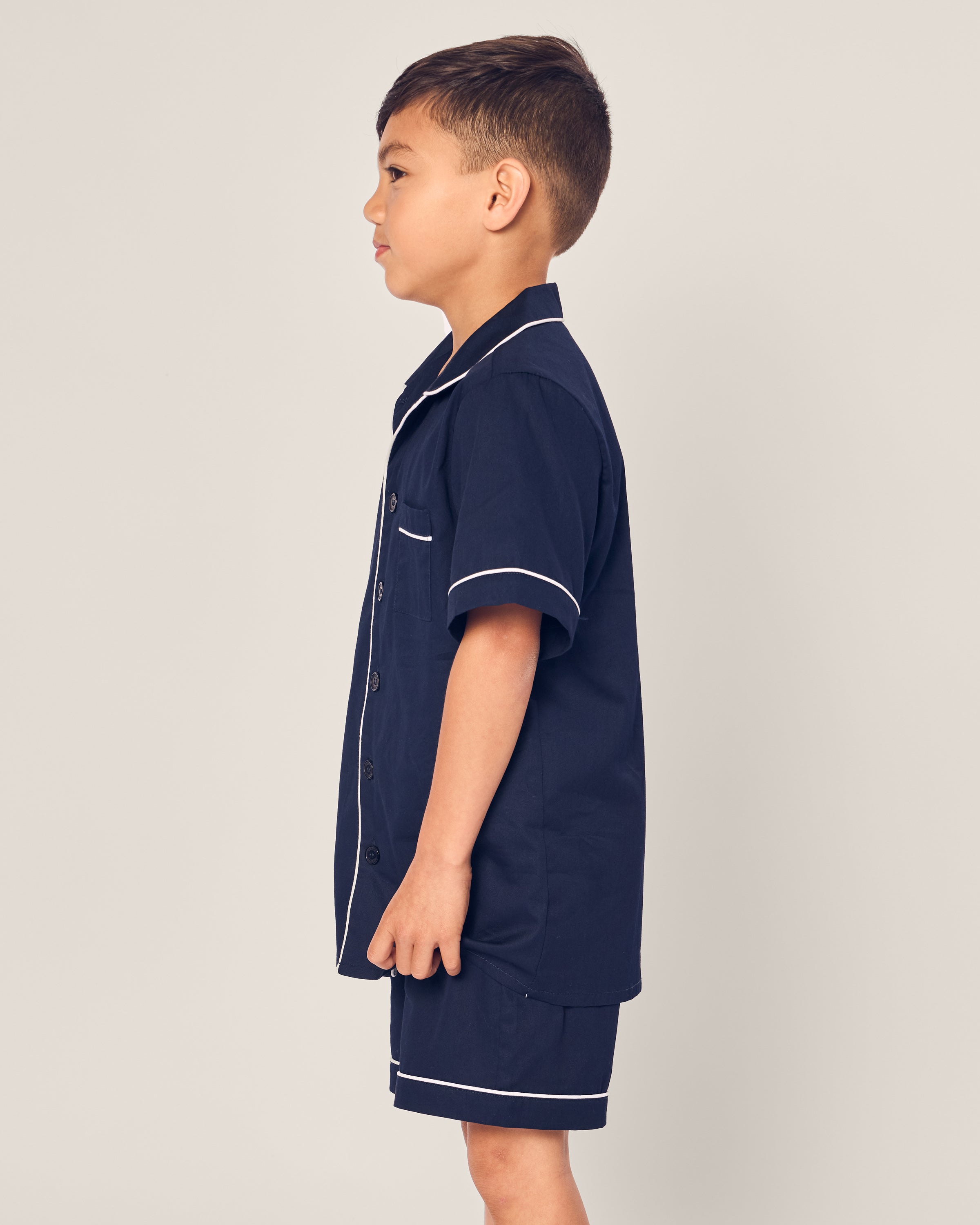 A young boy is in profile against a plain background, wearing Petite Plumes Kids Twill Short Sleeve Short Set in Navy, featuring white piping and crafted from inherently flame retardant fibers for luxurious sleepwear.