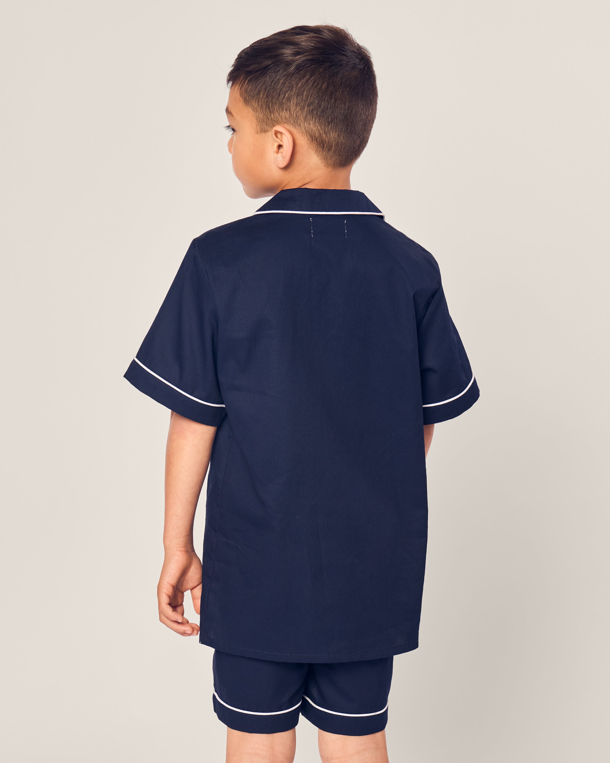 A boy stands facing away, wearing Petite Plumes Kids Twill Short Sleeve Short Set in Navy. His short brown hair complements the plain background, emphasizing the elegance of his inherently flame retardant fiber sleepwear.