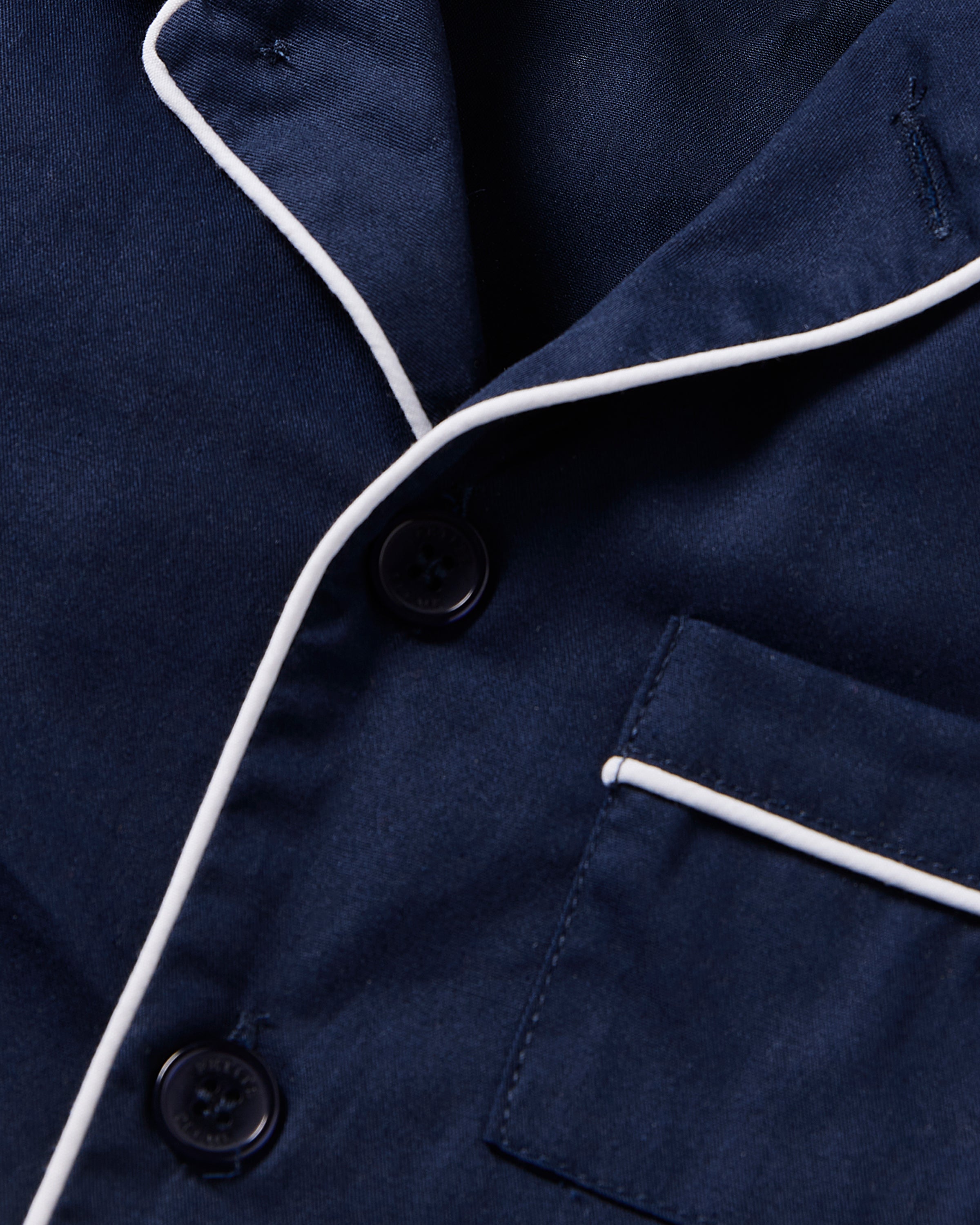 The close-up features Petite Plumes Kids Twill Short Sleeve Short Set in Navy, showcasing navy fabric with white piping, two black buttons, and a pocket accented by matching white trim, creating an elegantly refined sleepwear piece.