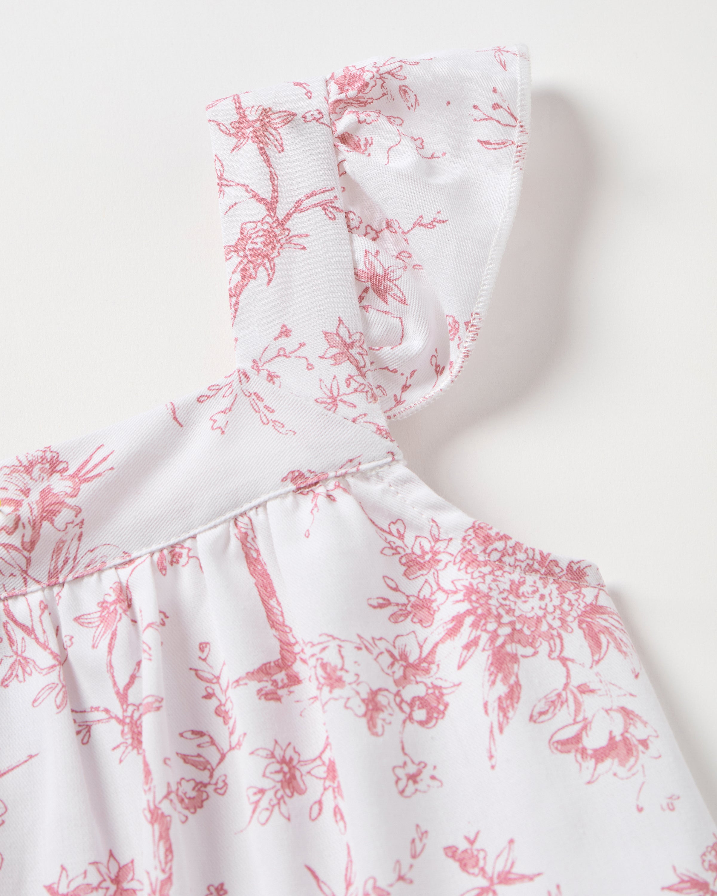 The Girls Twill Clara Short Set in Pink Timeless Toile by Petite Plume features delicate pink floral patterns on white fabric, a ruffled shoulder strap, and a gathered bodice, suggesting a feminine feel. The intricate design is evenly spread across the luxurious sleepwear piece.