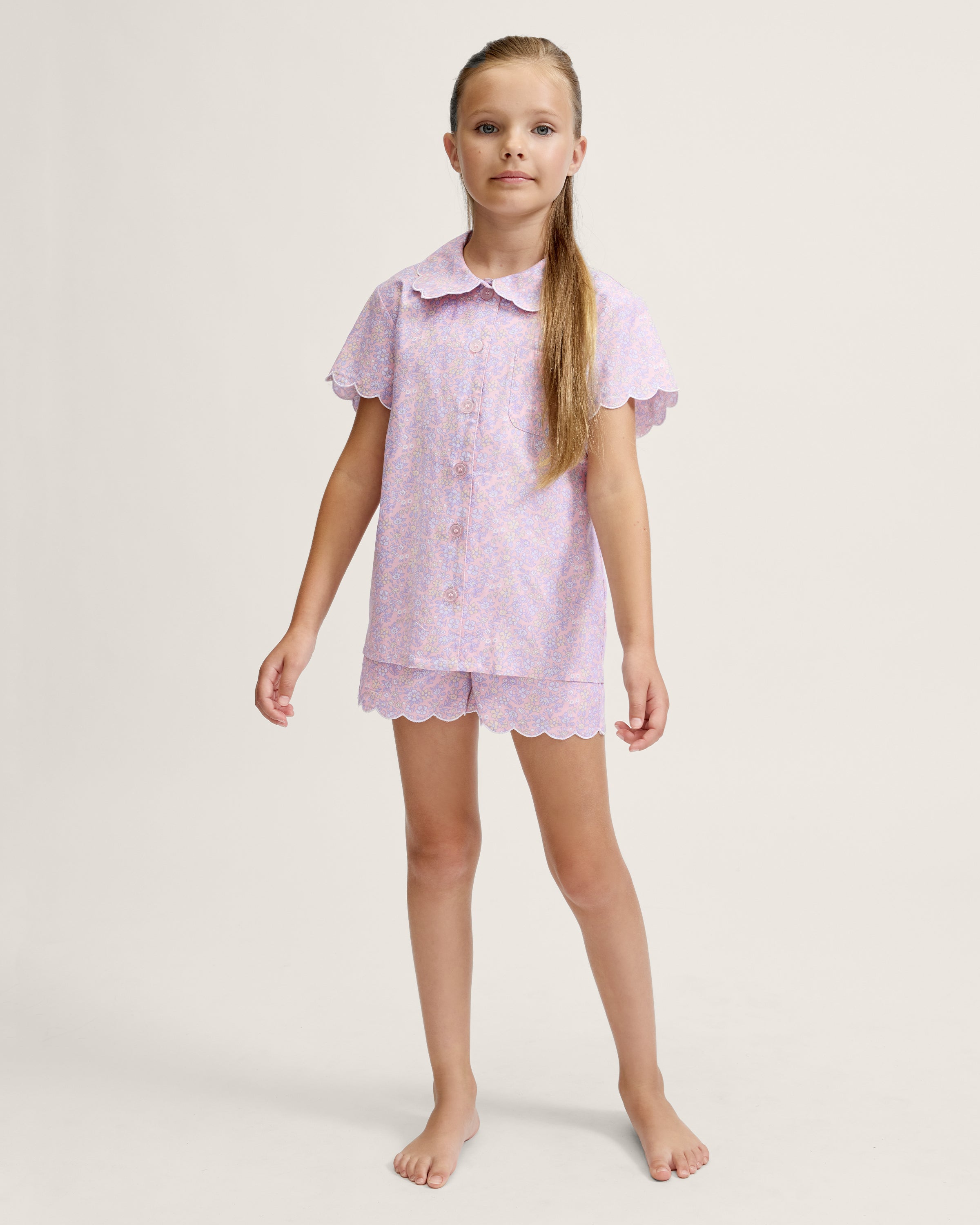 A young girl with long blonde hair stands facing forward, wearing the Kid's Twill Colette Short Set in Fleur Blush—an adorable pastel purple and pink pajama set with scalloped edges—against a plain light background.