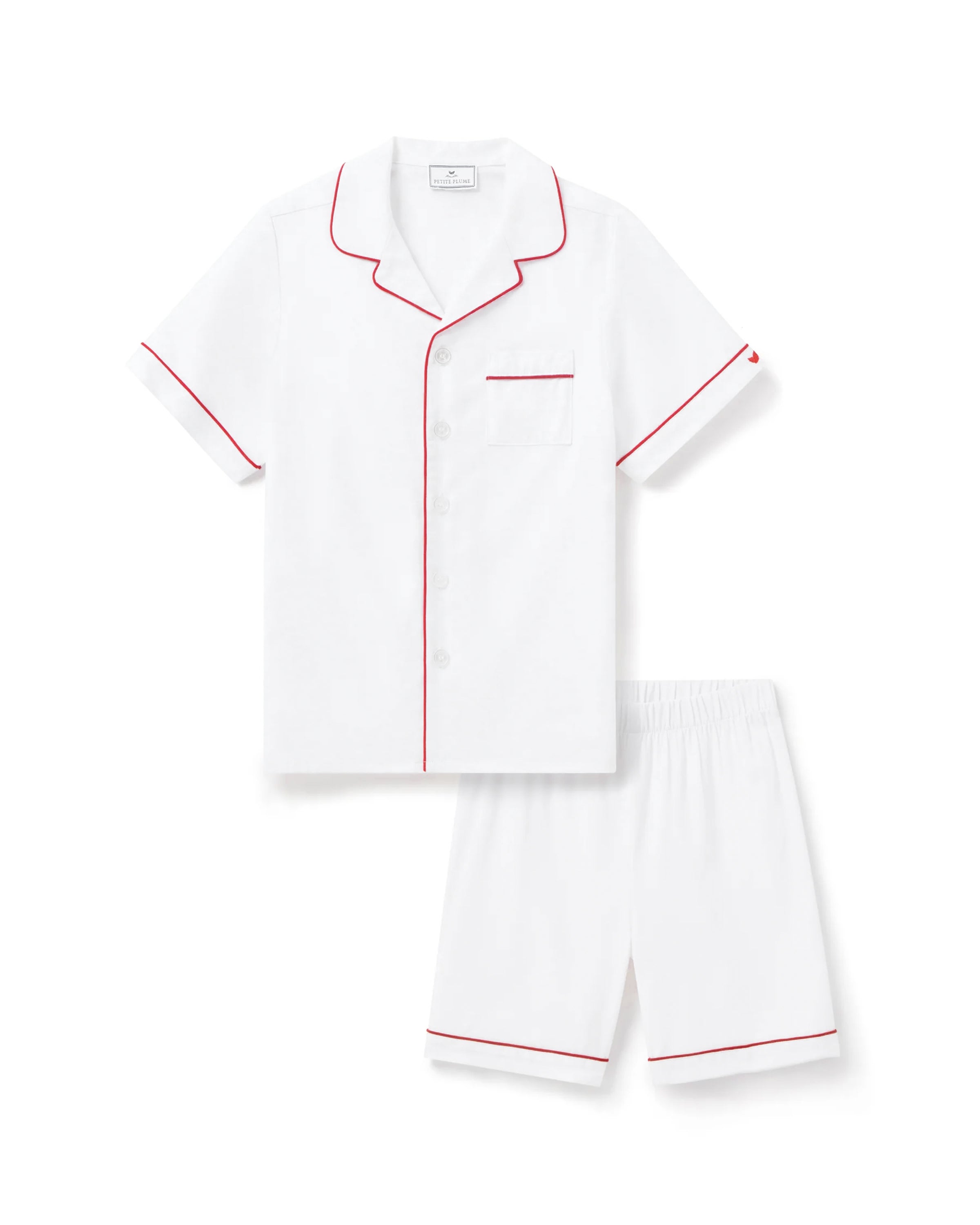 A child models Petite Plumes Kids White Twill Short Set with Red Piping, featuring a button-up short-sleeve shirt with a pocket and matching shorts. The flame-retardant sleepwear ensemble effortlessly combines style and safety against a plain background.