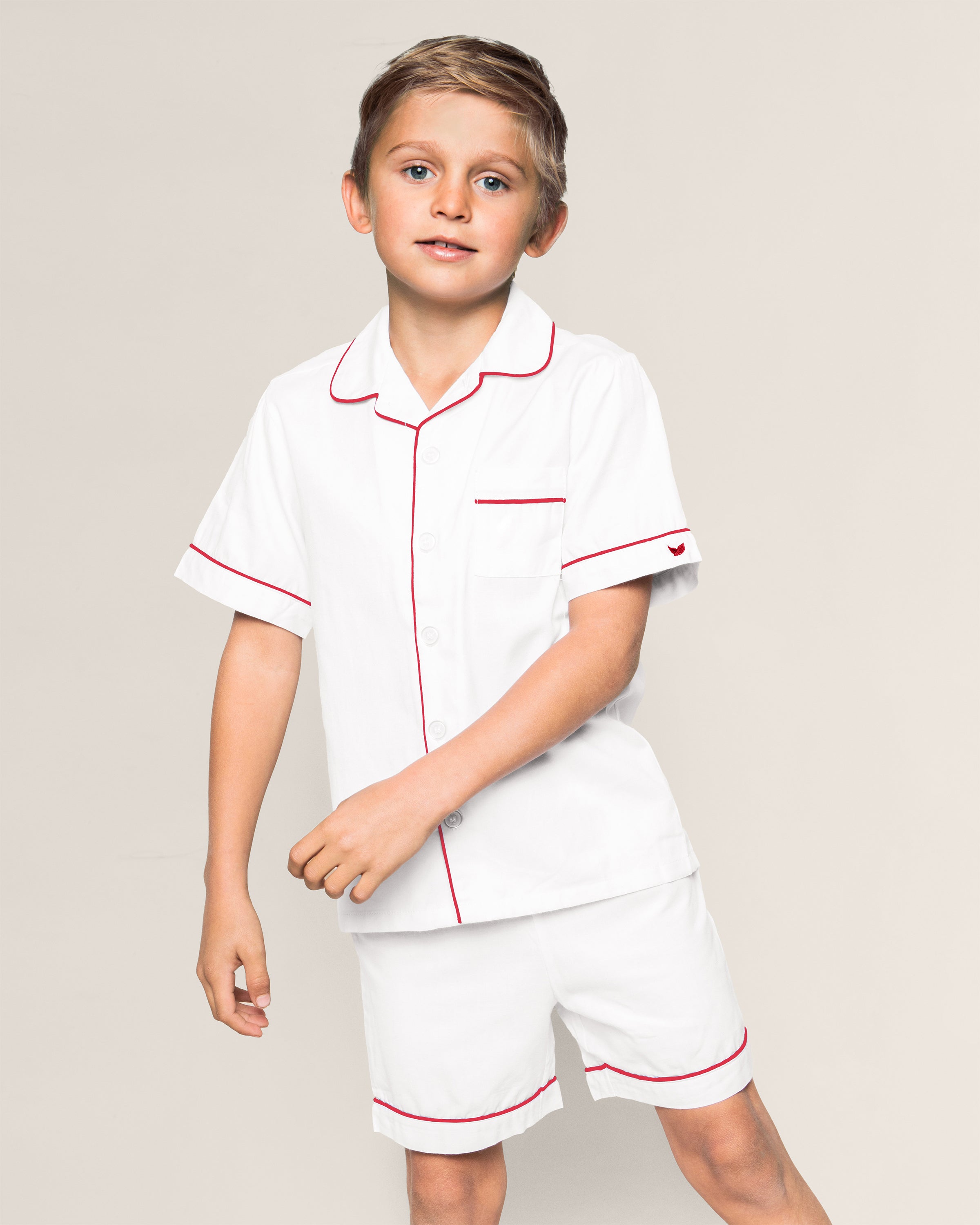 A child models Petite Plumes Kids White Twill Short Set with Red Piping, featuring a button-up short-sleeve shirt with a pocket and matching shorts. The flame-retardant sleepwear ensemble effortlessly combines style and safety against a plain background.
